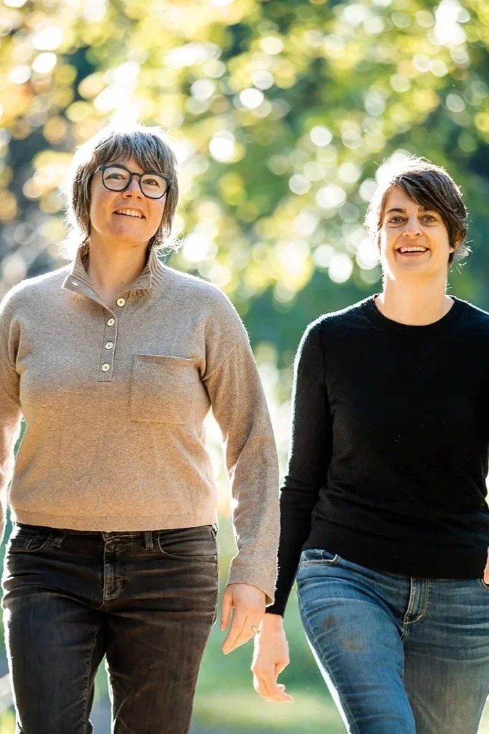 Two women walking outdoors on a sunny day, smiling. One woman has short brown hair, glasses, and is wearing a beige sweater and dark jeans. The other has short dark hair and is wearing a black sweater and blue jeans. They are surrounded by green trees and bokeh sunlight.