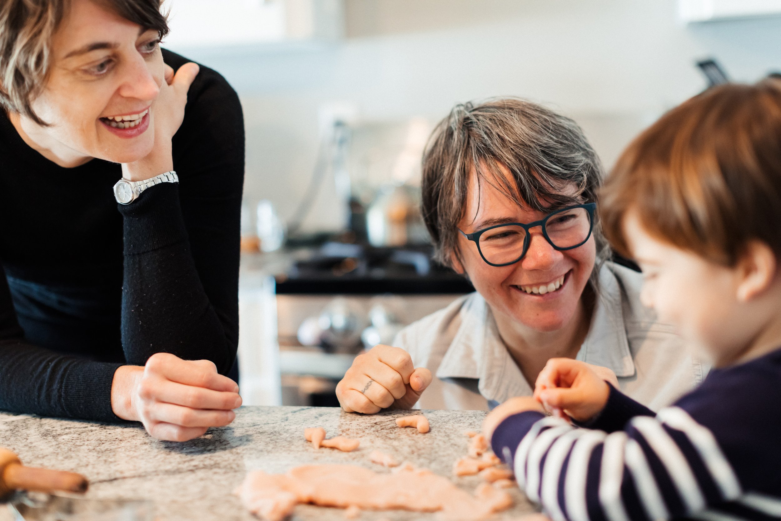 Three people, two adults and one child, smiling and playing with clay on a kitchen countertop.