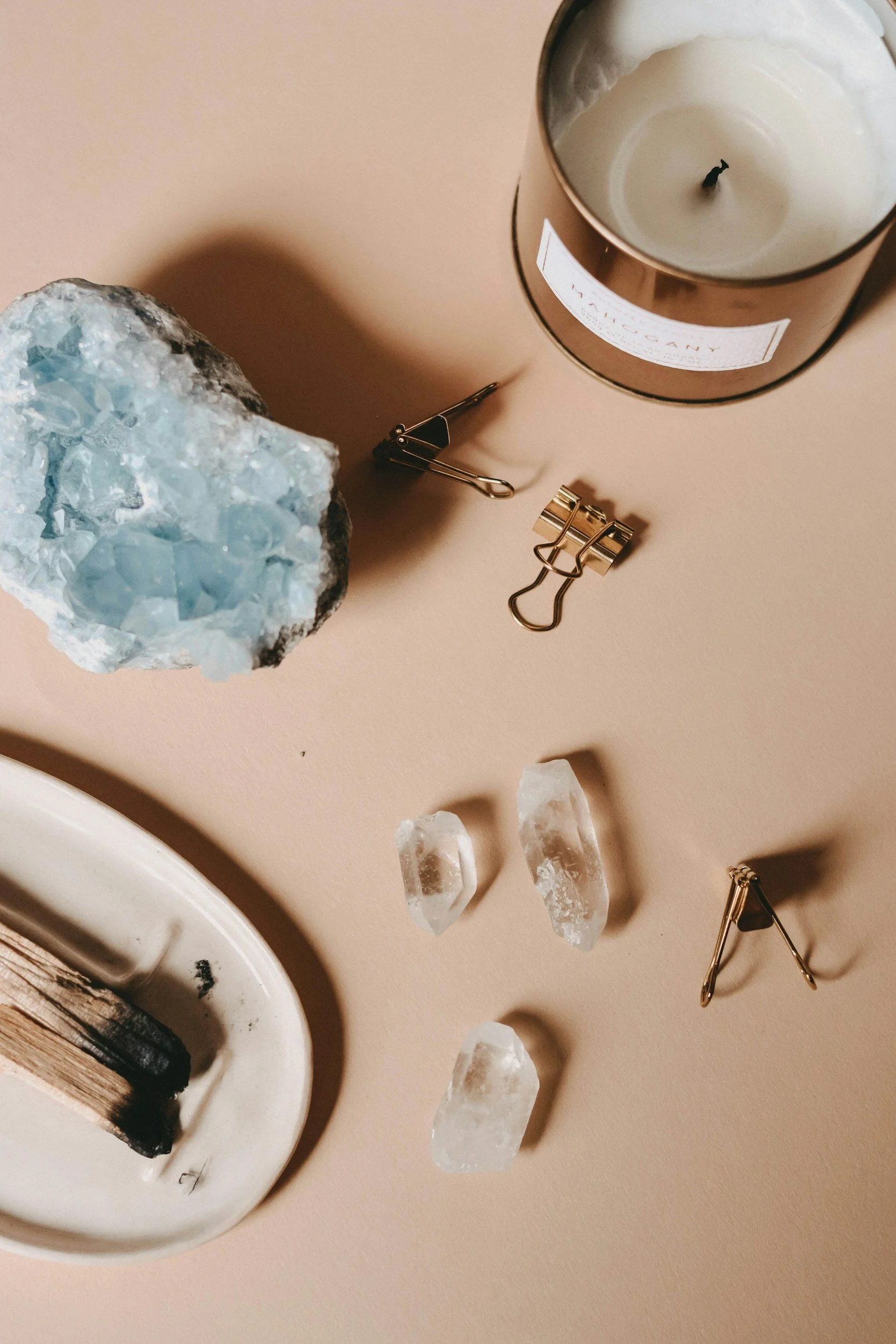 A flat lay of a partially burned candle, a large light blue mineral or geode, several small clear quartz crystals, a small brass binder clip, a safety pin, and a small brass triangle on a neutral background.