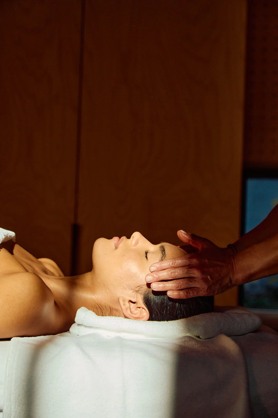 A woman receiving Reiki and is lying on her back on a massage table, with a towel covering her lower body, while a therapist holds her forehead with both hands.