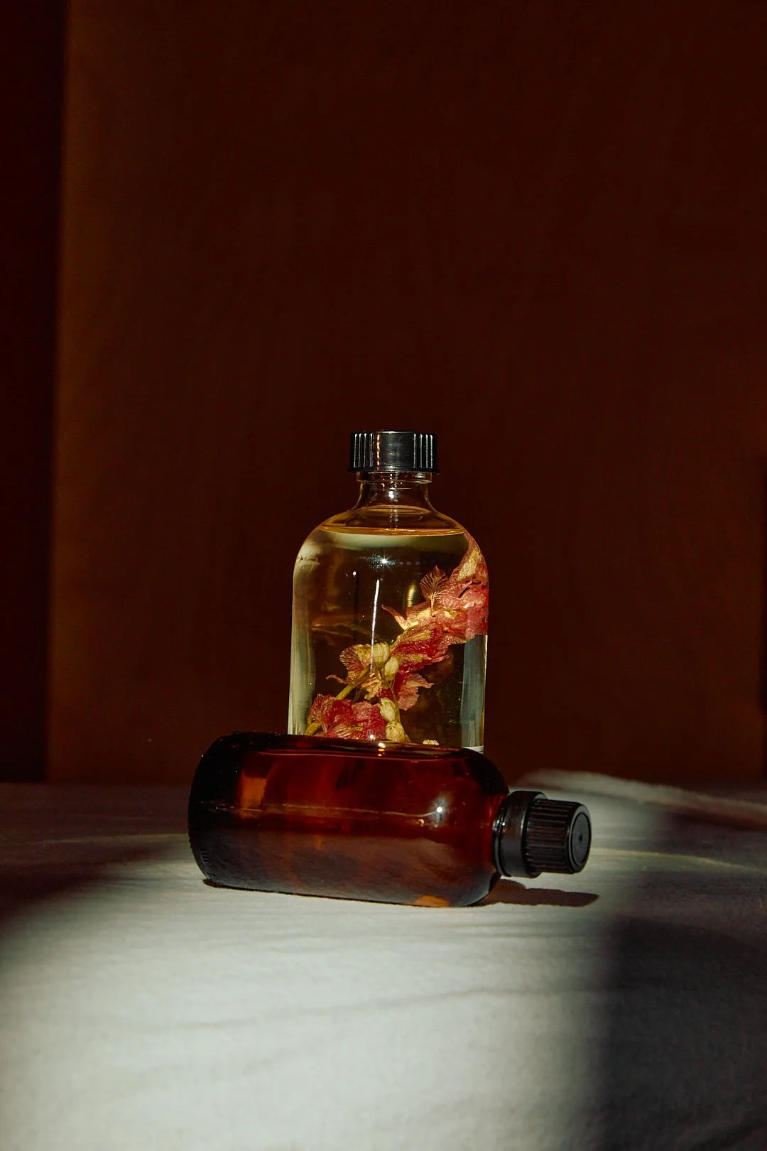 Two bottles, one with a dark amber color lying on its side and the other with a clear liquid and a plant submerged inside, standing upright on a surface with a dark background.
