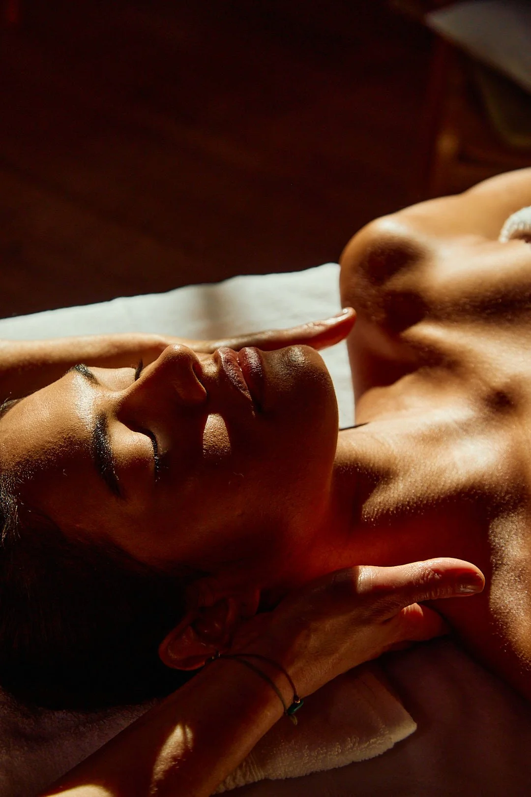 A woman receiving a head massage on her back, lying on a massage table, with warm lighting highlighting her face and upper body.