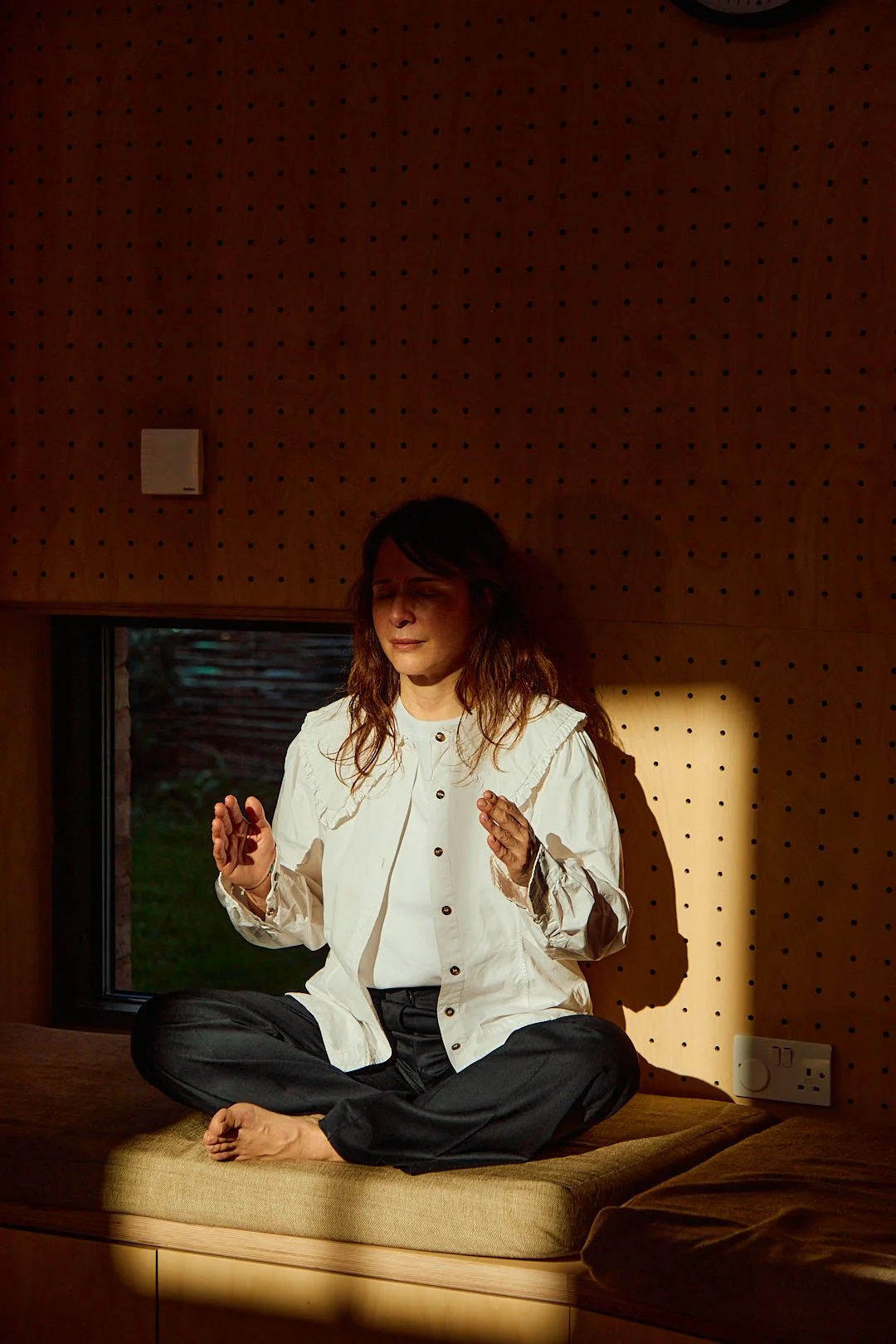 A woman with long dark hair sitting cross-legged on a cushion inside a wooden room, meditating with her eyes closed as sunlight casts a shadow on the wall behind her.