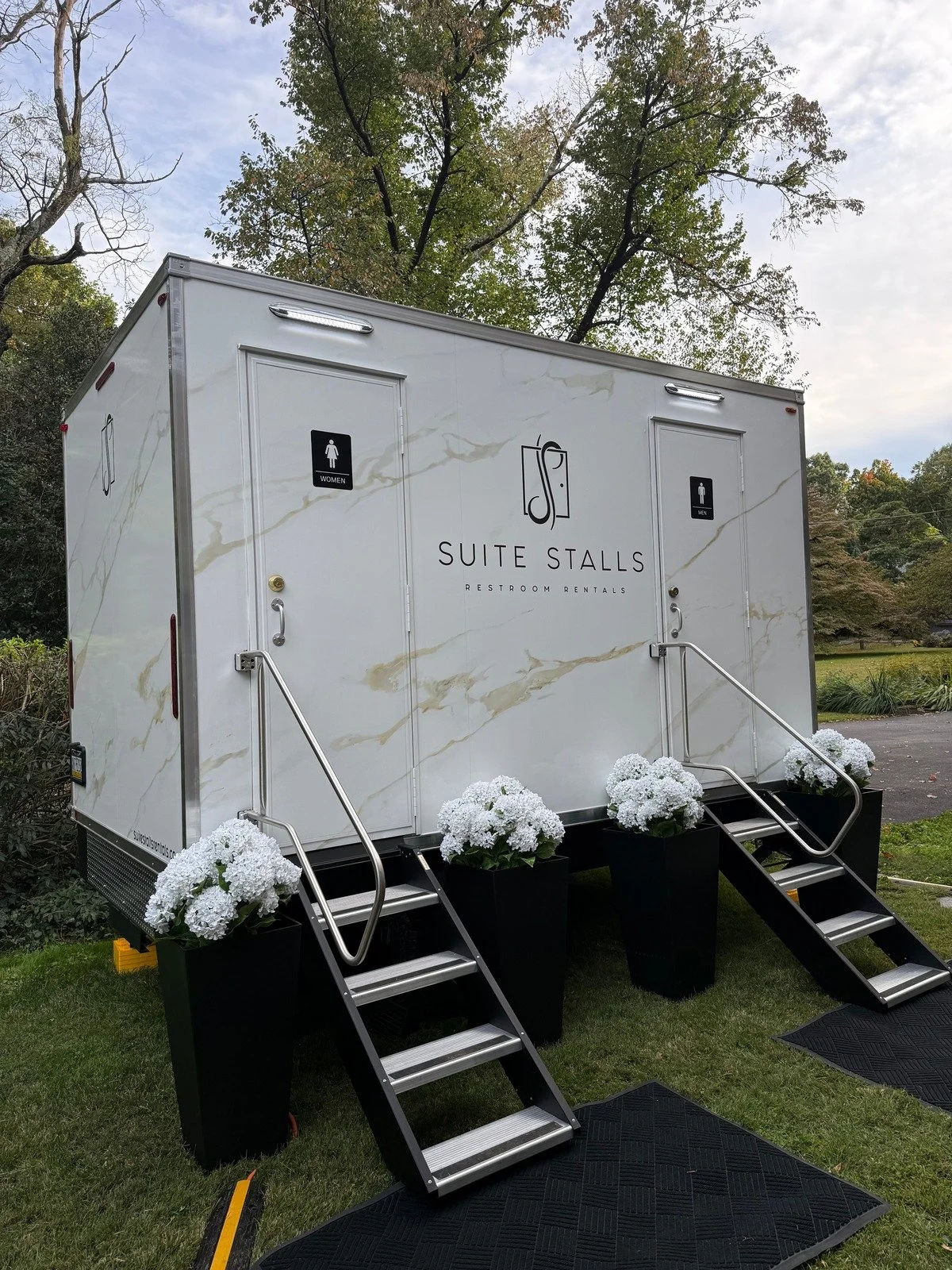 Suite Stalls restroom trailer with white flowers in black planters, set up on a lawn for an outdoor wedding.