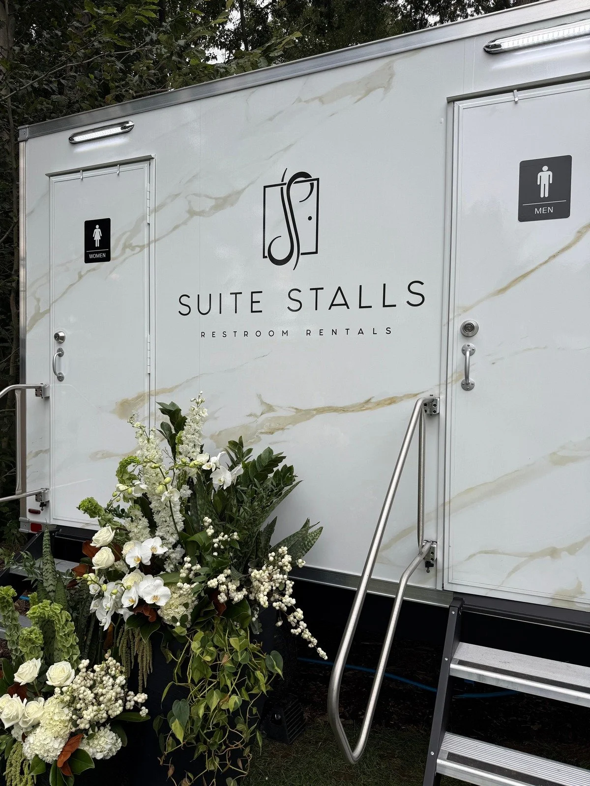 White flowers and greenery beside the Suite Stalls trailer entrance at a formal outdoor event.