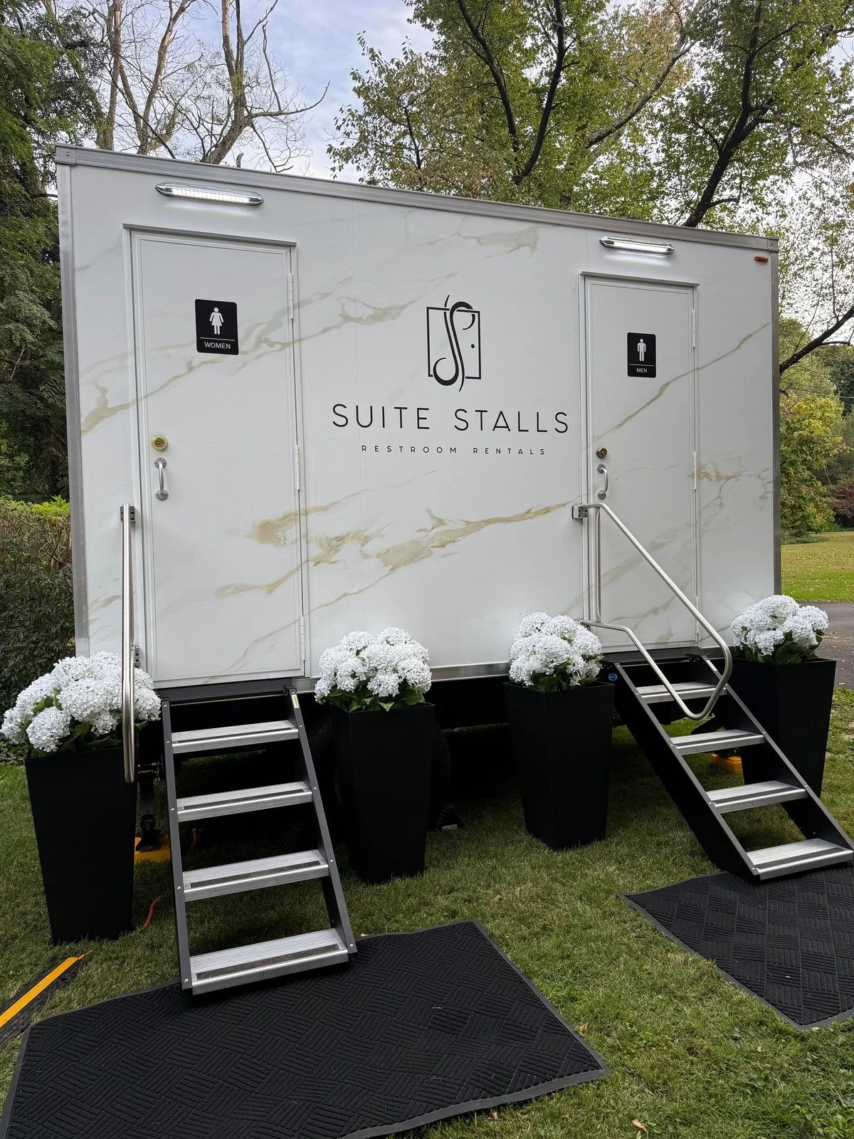 Suite Stalls restroom trailer with white floral arrangements and black steps at an outdoor wedding.