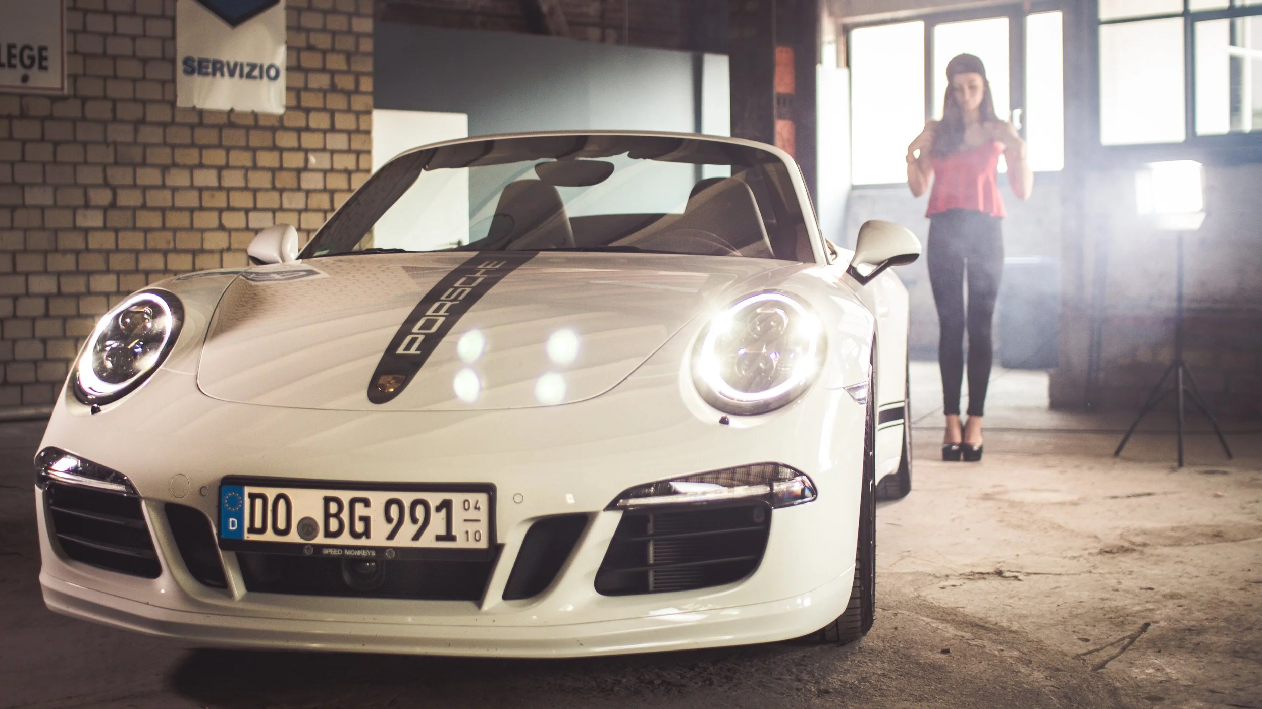 Weiße Porsche-Auto im Parkhaus, im Hintergrund eine Frau mit roten Top und schwarzen Hosen, Frontansicht des Autos, mit Porsche-Logo auf der schwarzen Markierung auf der Motorhaube, deutsches Nummernschild.