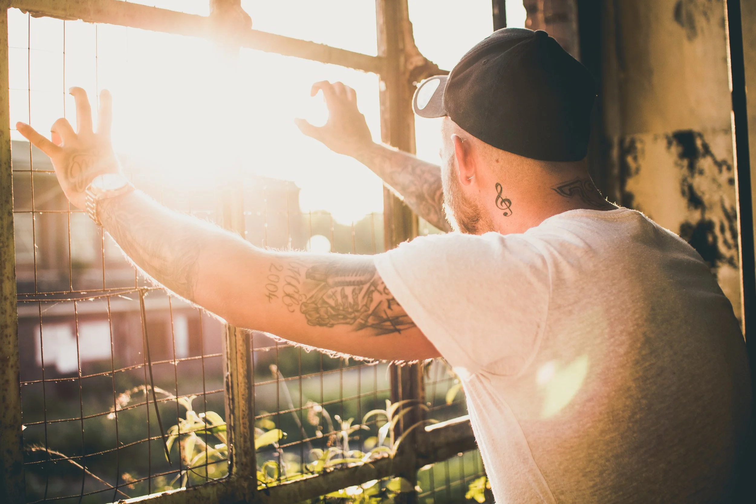 Ein Mann mit Tattoos in einem weißen T-Shirt trägt eine schwarze Kappe und steht an einem Fenster, während die Sonne scheint.