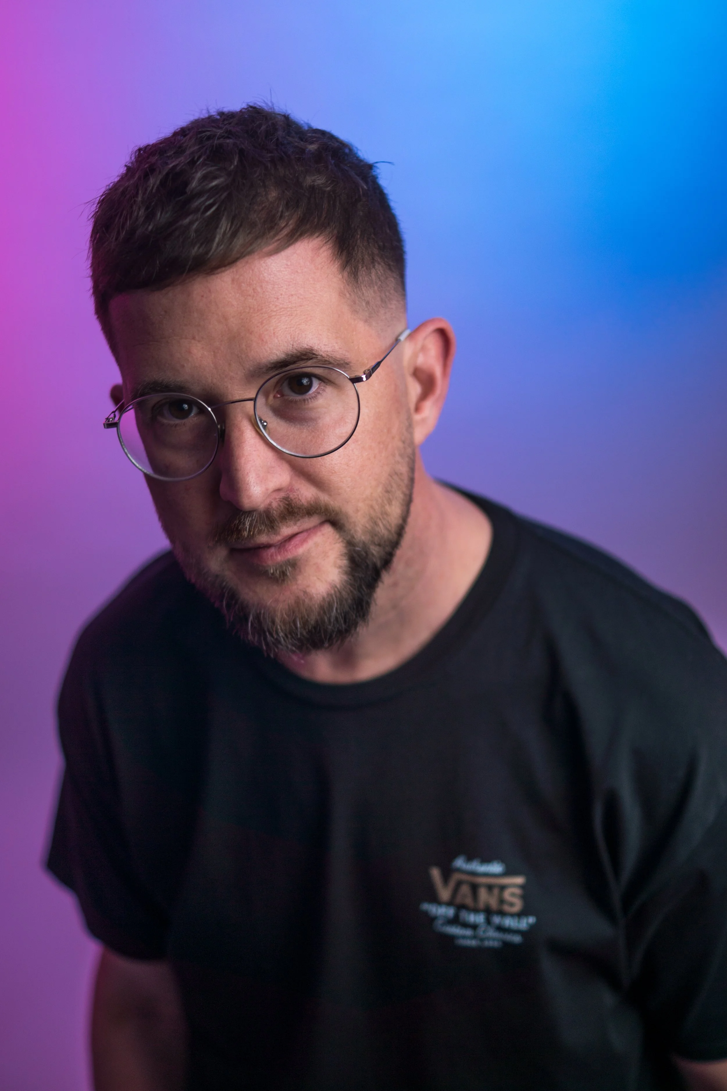 Junge Mann trägt schwarze Vans-T-Shirt, trägt runde Brille, hat kurzen braunen Haaren, trinkt Bier, professionelles Porträt vor farbigem Hintergrund