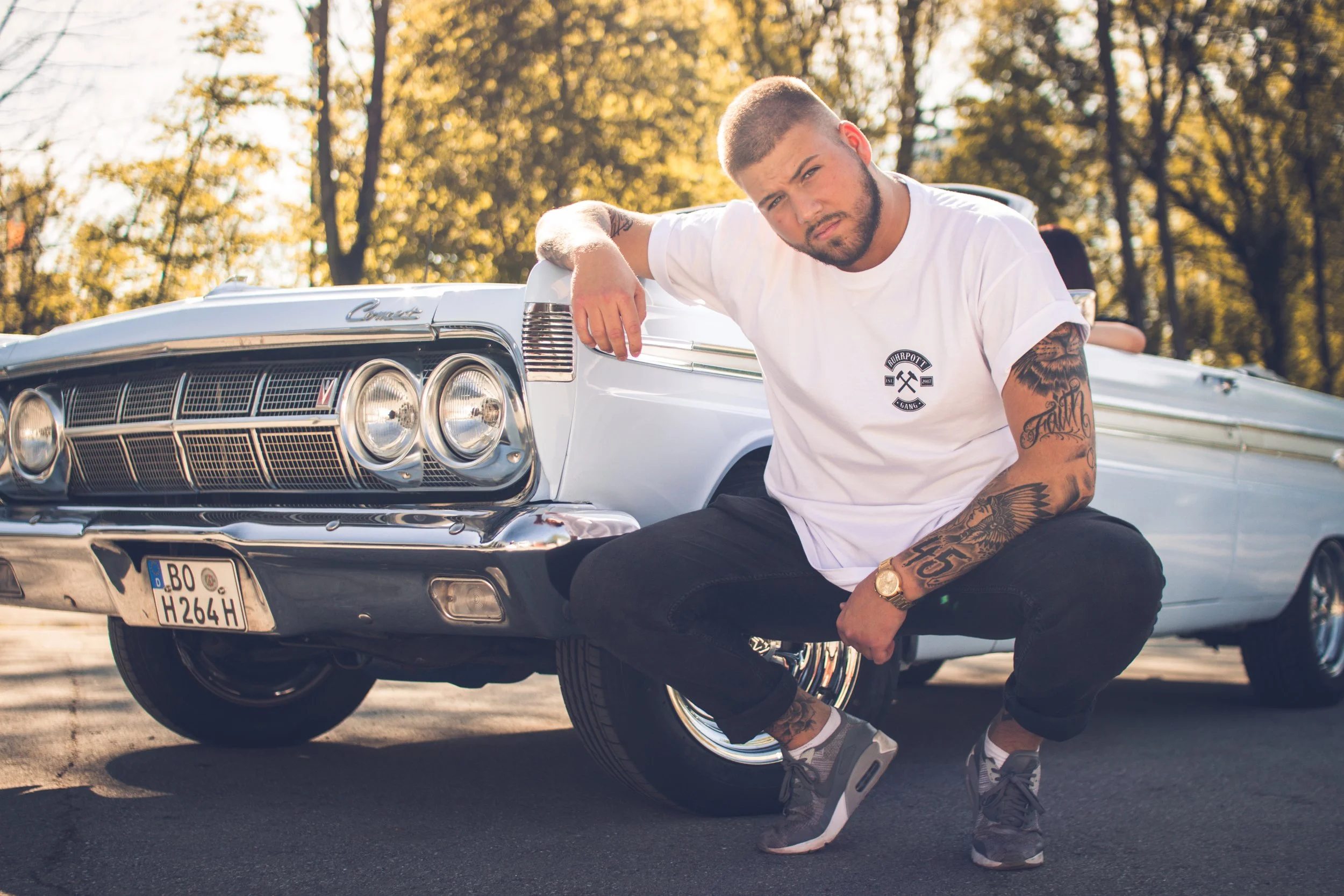 Junger Mann mit Tattoos vor einem weißen Oldtimer, im Freien bei Sonnenuntergang