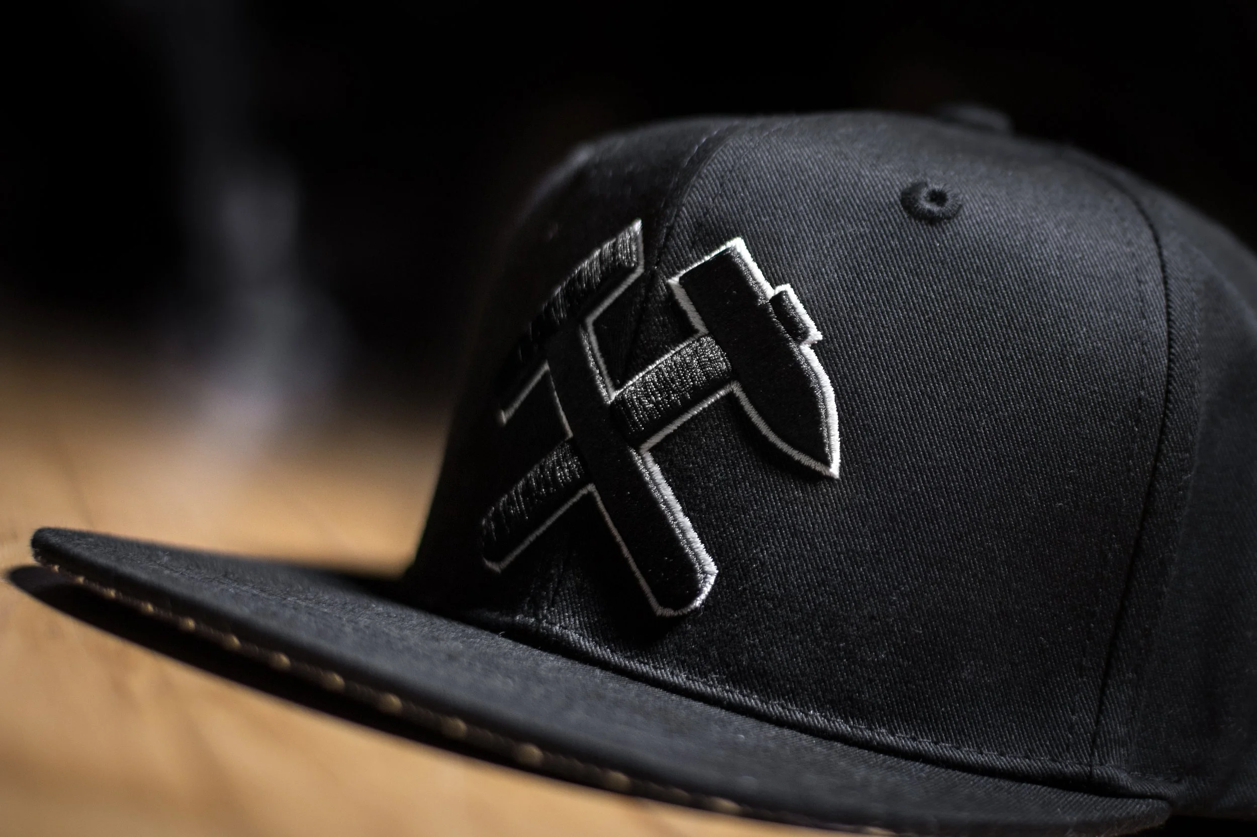 Schwarze Baseballkappe mit gesticktem Logo, liegend auf einer Holzoberfläche.