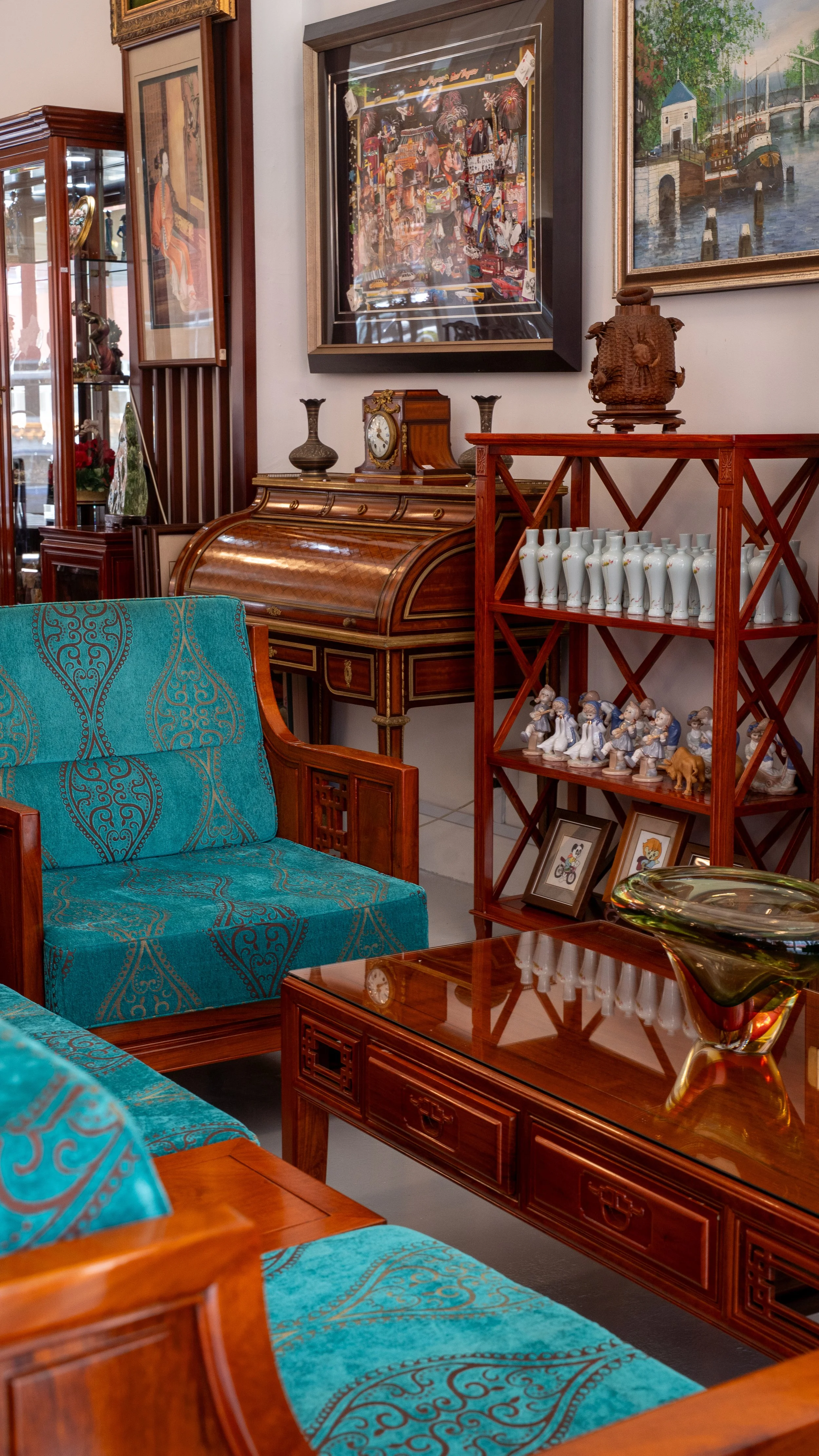 Vintage living room with wooden furniture, including a turquoise upholstered sofa and armchair, a polished wooden coffee table, a wooden secretary desk with a clock and vases, a wooden shelving unit with porcelain figurines and framed photos, and framed artwork on the walls featuring landscapes and abstract scenes.