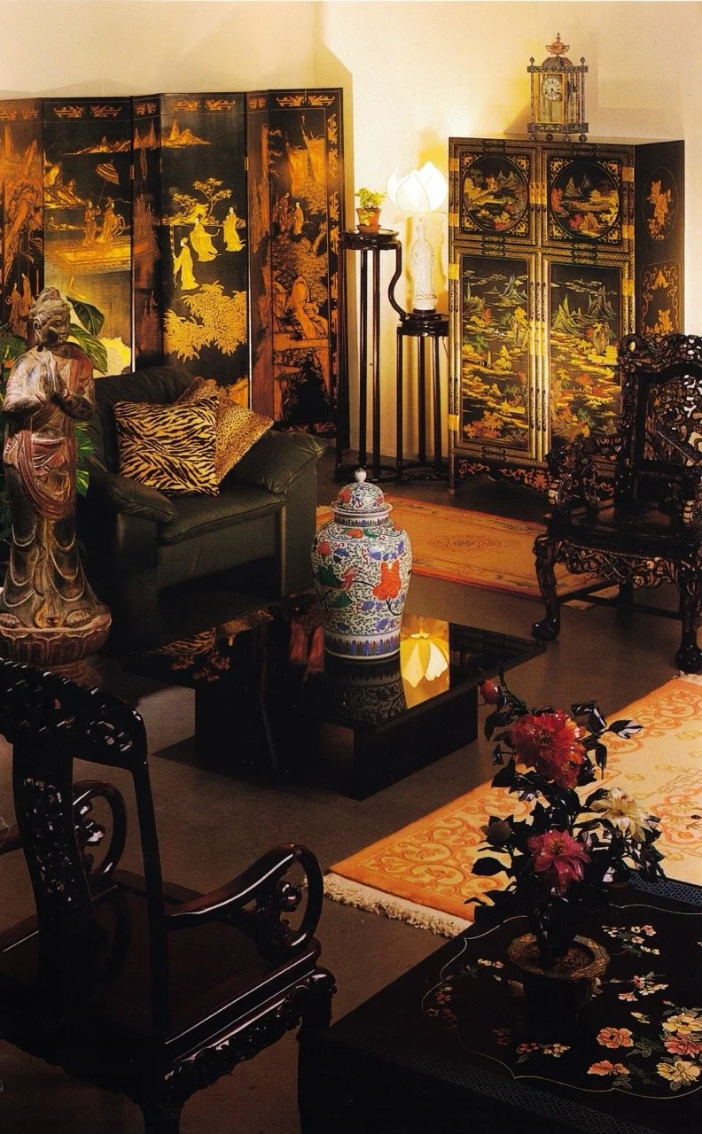 Traditional Asian-inspired living room with black and gold Asian folding screen, black decorative cabinet with landscape painting, small table with a potted plant, standing lamp, ornate wooden chairs, floral-patterned vase, and decorative elements.