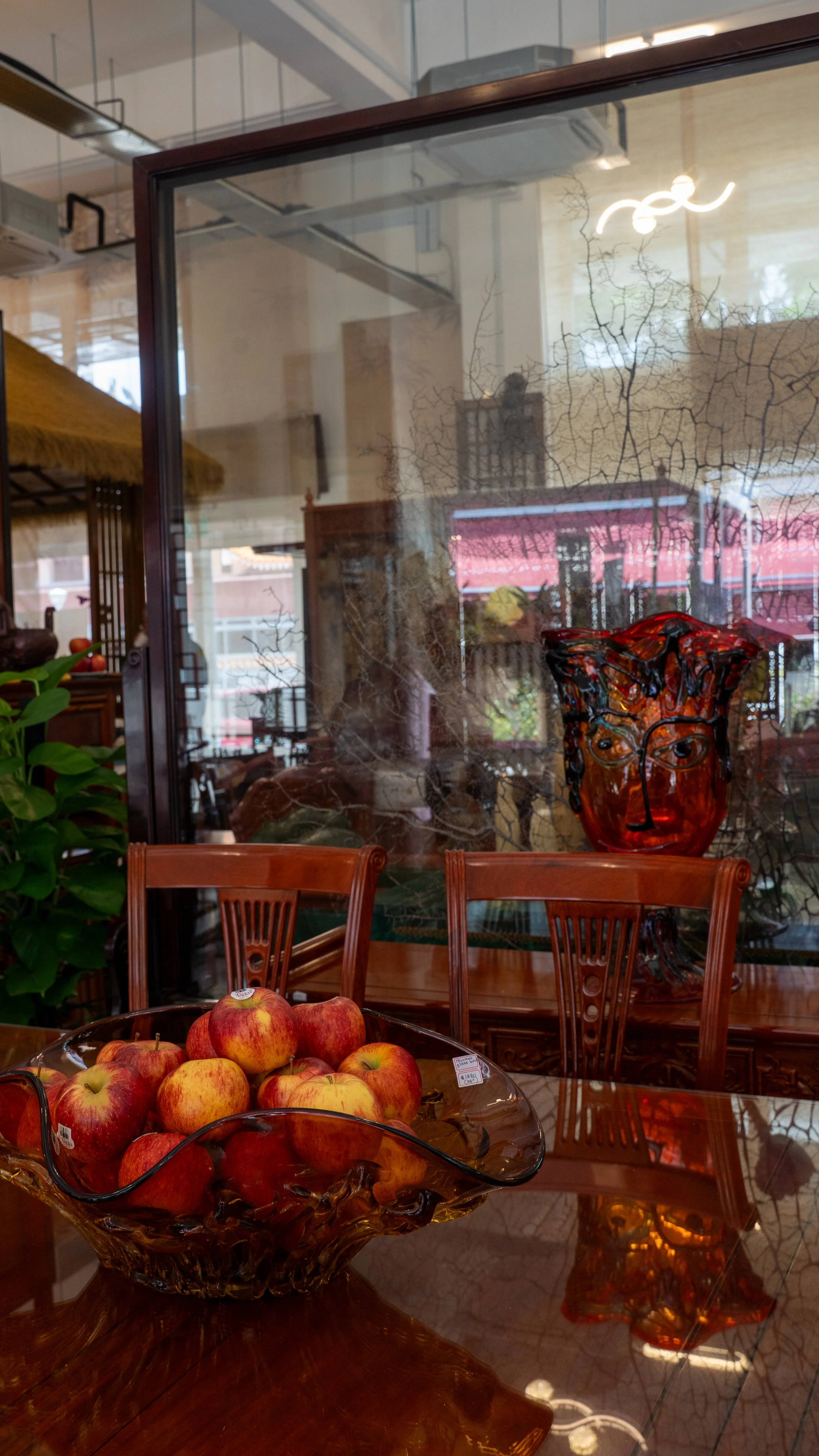 Display of red apples in a decorative glass bowl on a polished wooden table in an indoor setting with a wooden chair, glass sculpture of a face, and reflections of the room.