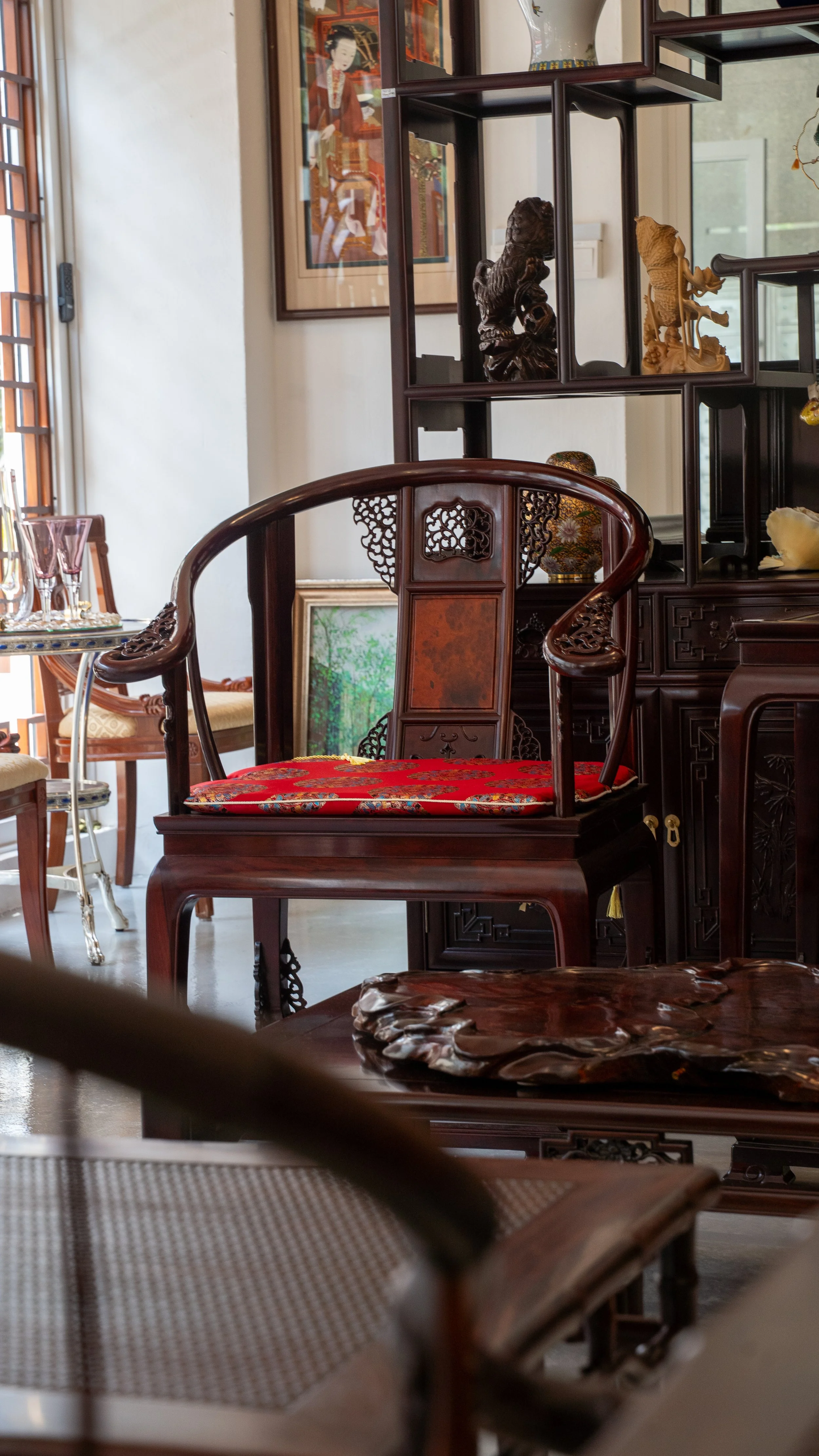 Interior of a room with antique Asian-style furniture, including a carved wooden chair with a red cushion, a shelf with decorative figurines, and framed artwork on the wall.