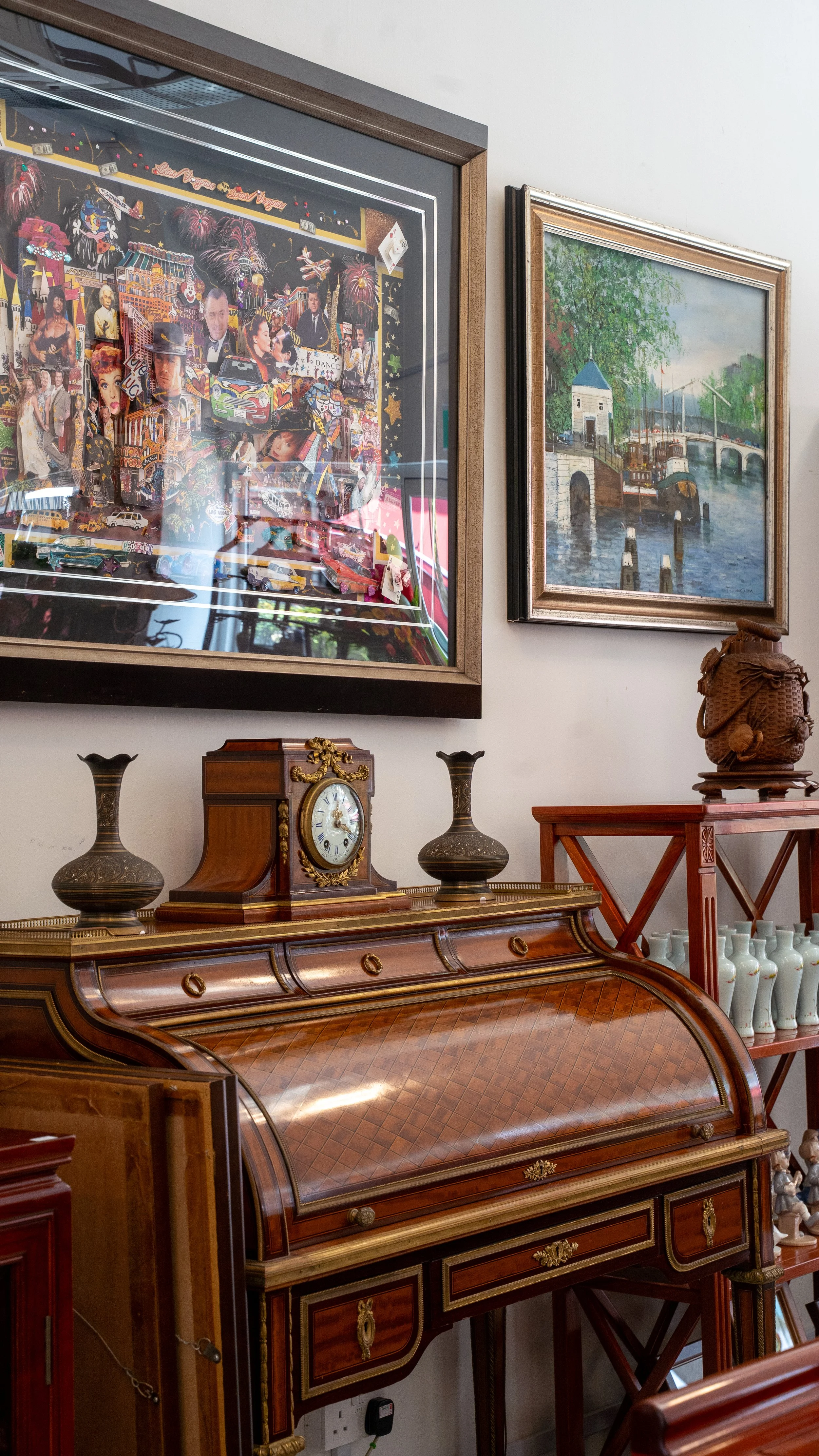 A corner of a room with two framed paintings on the wall and antique wooden furniture, including a clock and decorative vases.