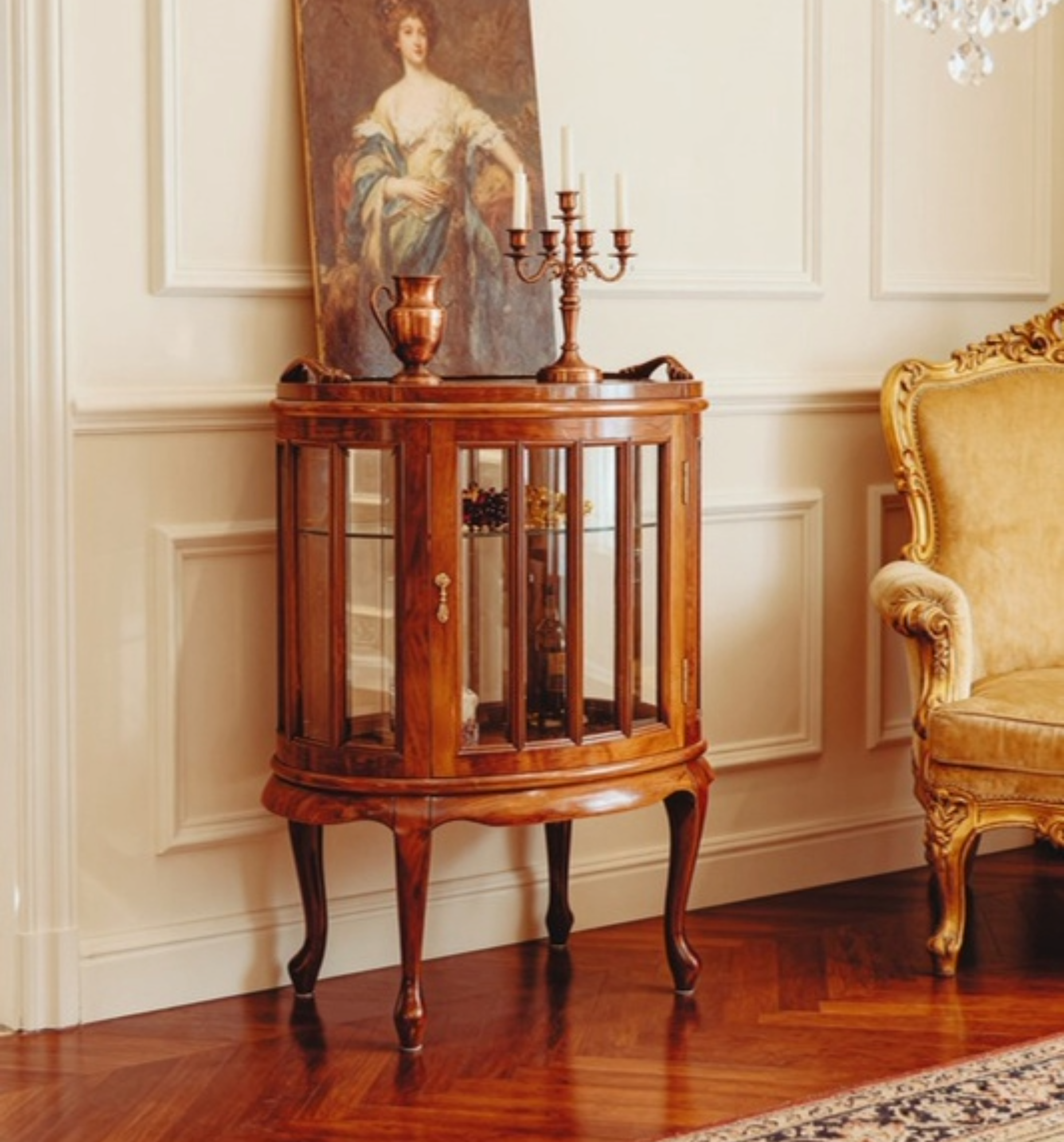 A vintage wooden display cabinet with glass panels and curved legs, decorated with a bronze candlestick holder and a small bronze pitcher, located in an elegant room with wood flooring, a traditional painting of a woman in period clothing, and a yellow upholstered armchair.
