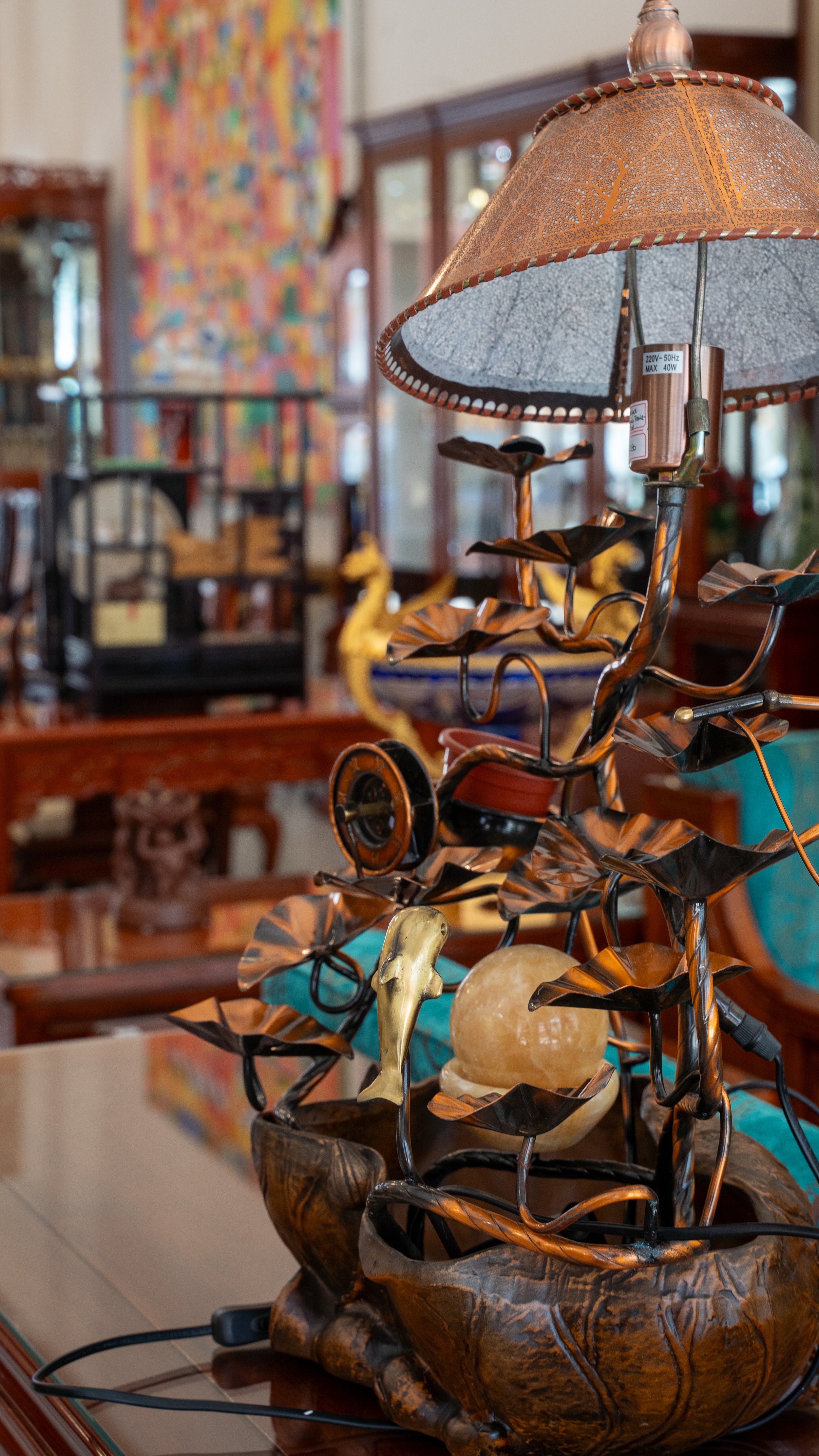 Decorative table lamp with metal leaves, a sphere, and a lamp shade in a room with wooden furniture and colorful artwork in the background.
