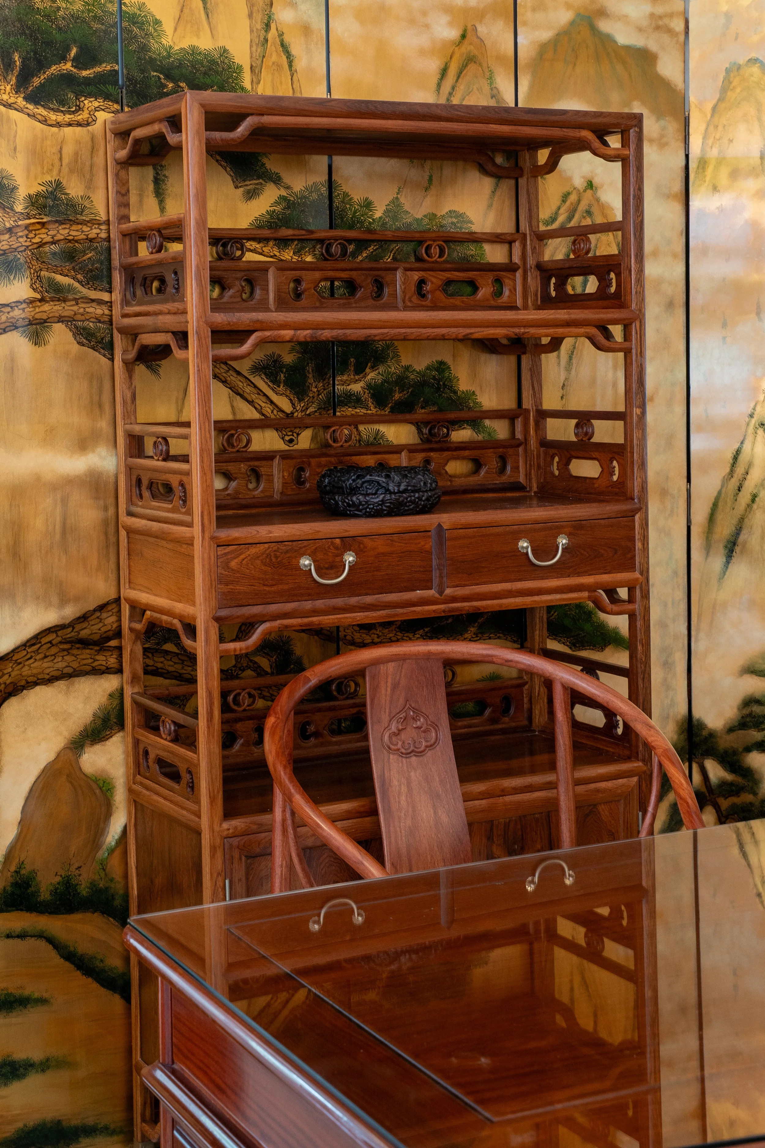 A wooden display shelf with three tiers and a drawer, sitting in front of a traditional Asian-style painted wall. There is a carved black object on the middle shelf and a wooden chair with an ornate backrest in front of the shelf. A glass-topped table is partially visible in the foreground.