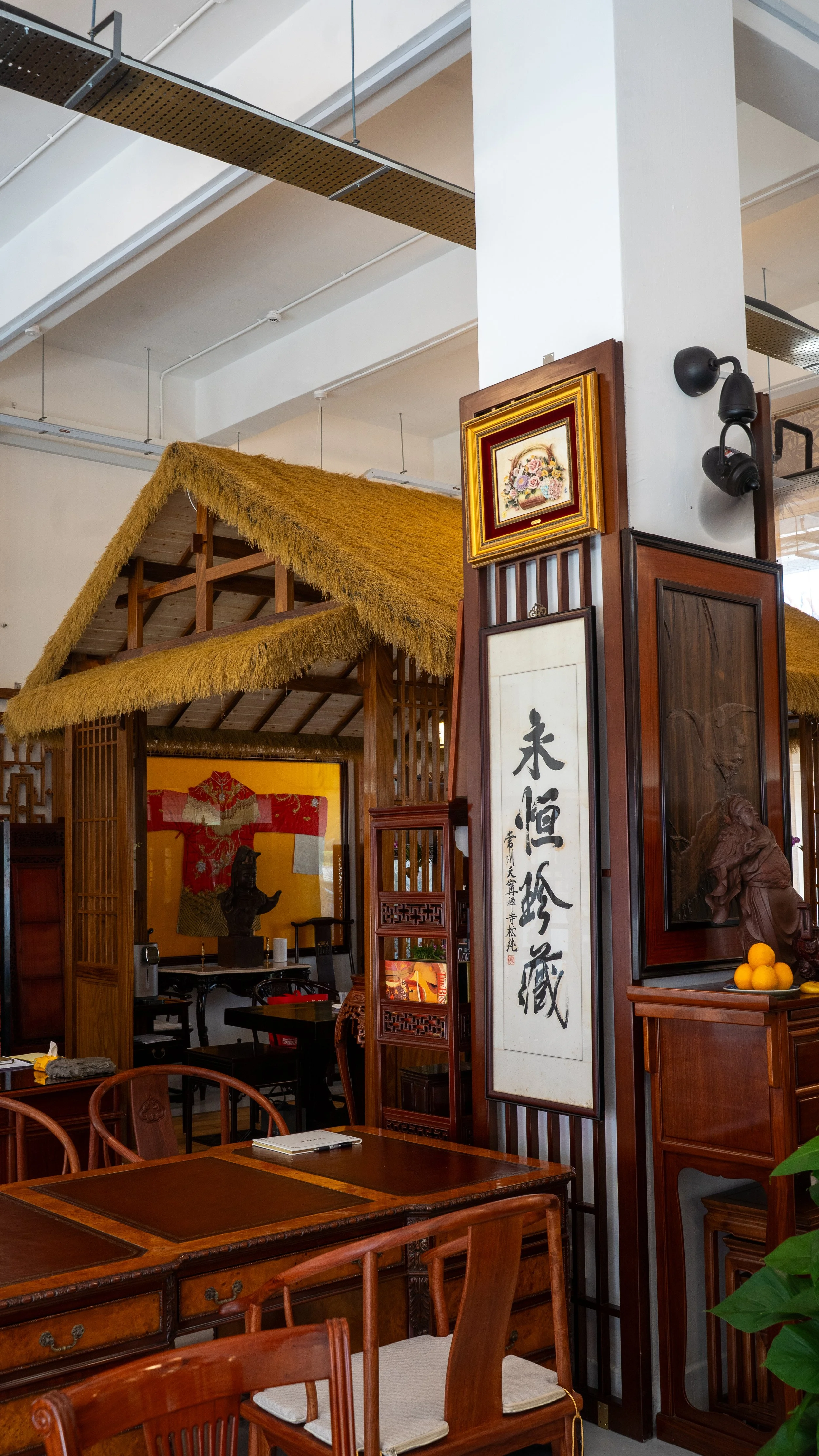 Interior of a restaurant decorated with Asian-inspired furniture and artwork, including Chinese calligraphy, wooden tables and chairs, and a decorative thatched roof structure.