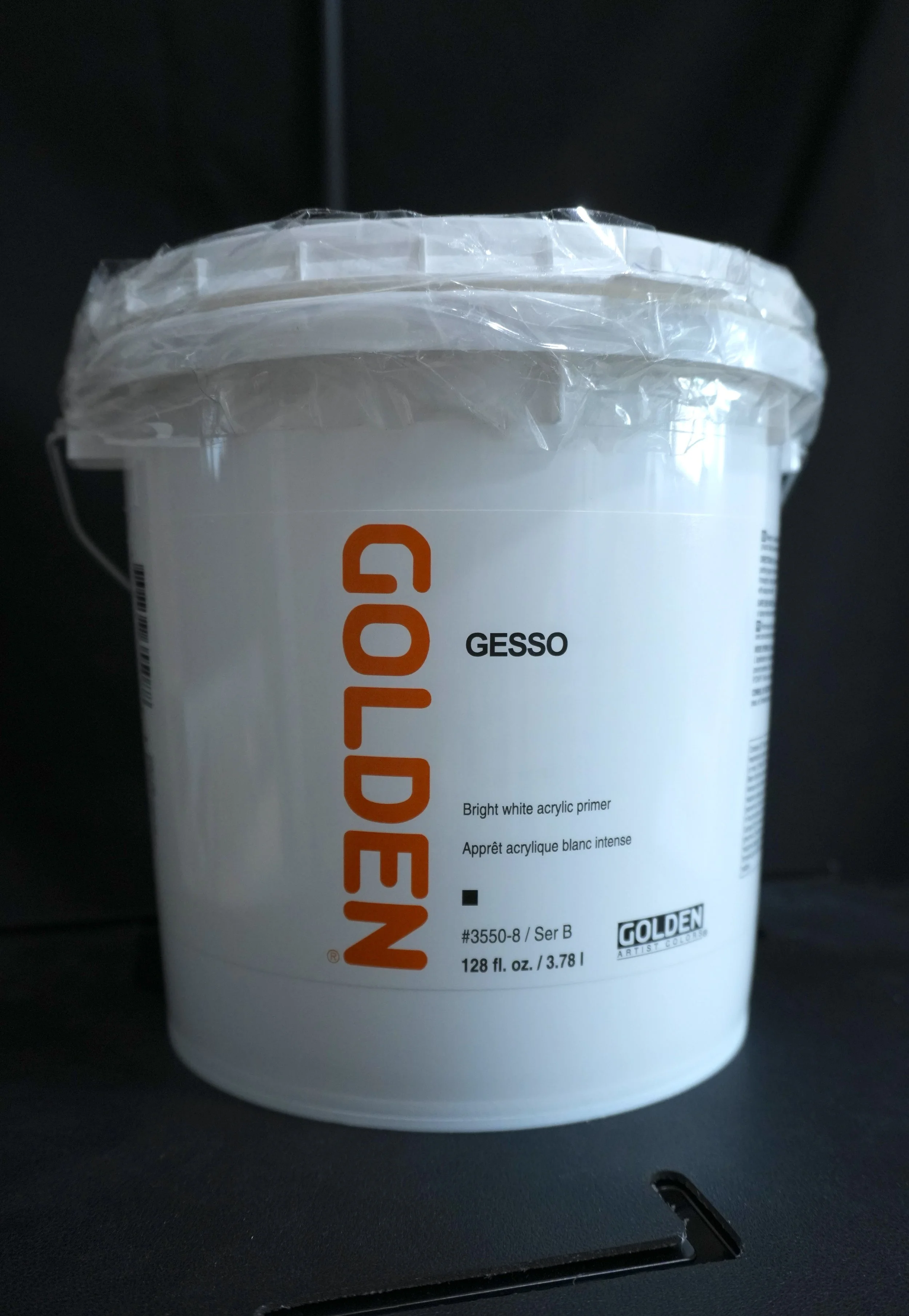 A white bucket of Golden Gesso, a bright white acrylic primer, with a plastic seal on top and a black background.