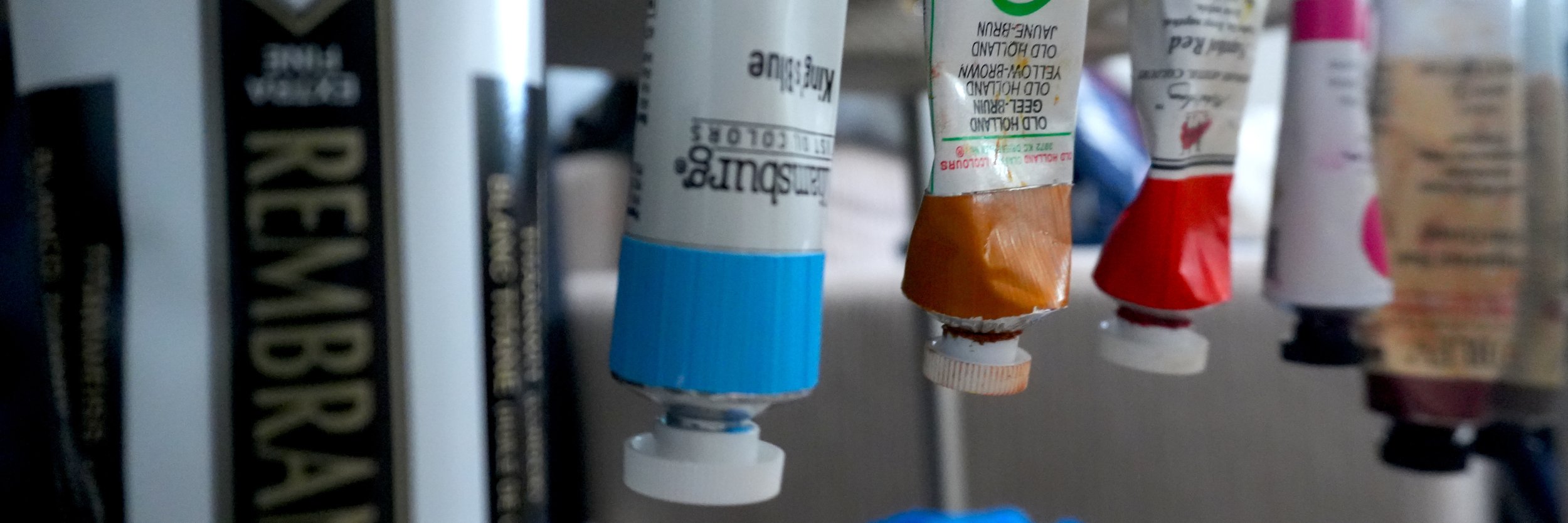 Close-up of various tubes of acrylic paint hanging upside down, with visible caps and labels, in an art or painting supply setting.