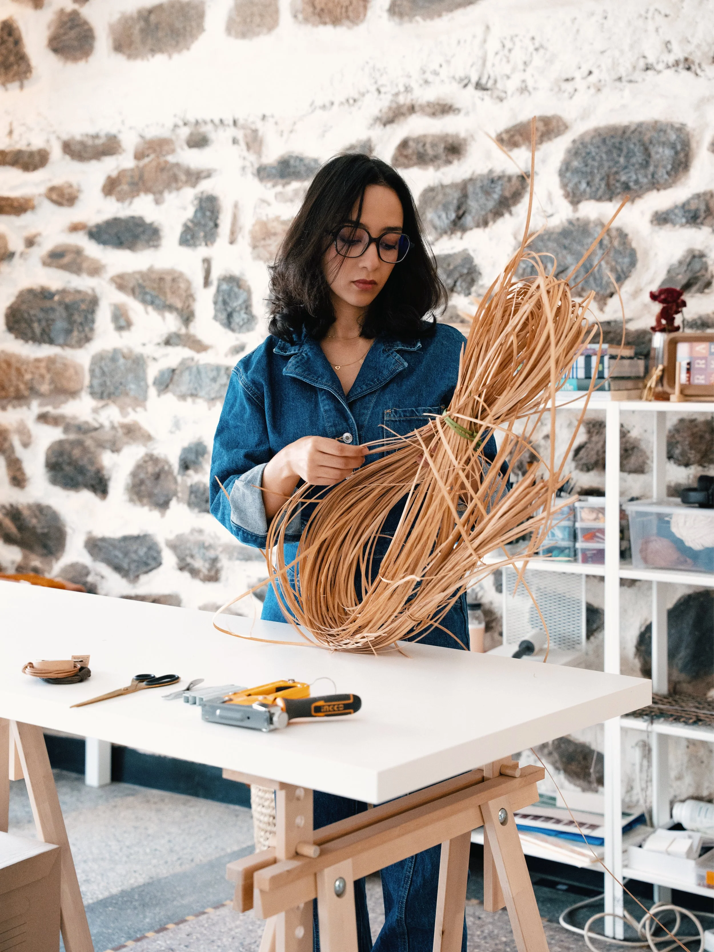 Young woman with glasses and dark hair working on a craft project with dried palm leaves at a white table in a room with a stone wall.
