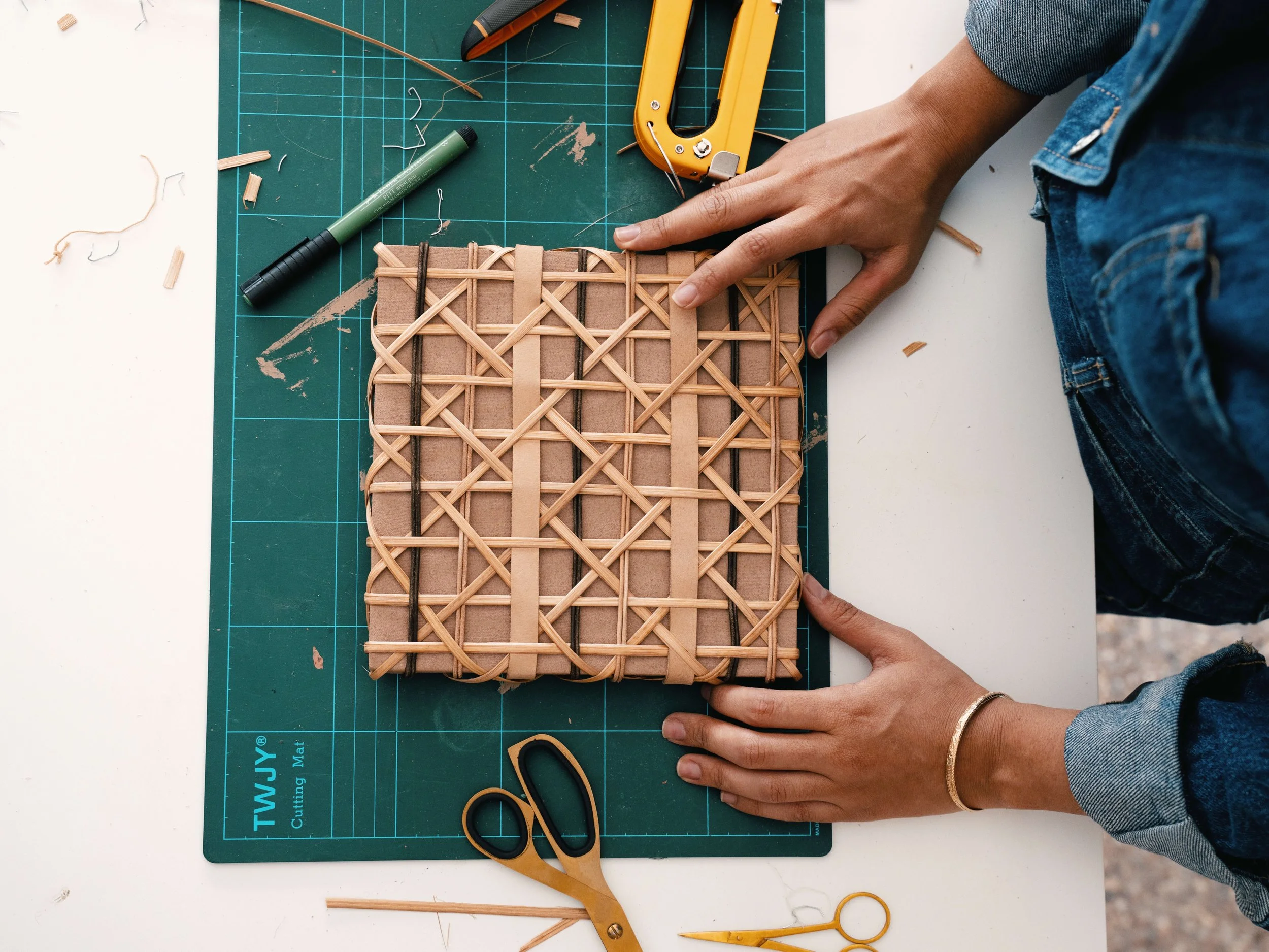 Person working on a woven basket with a cutting mat, scissors, and crafting tools on a white surface.