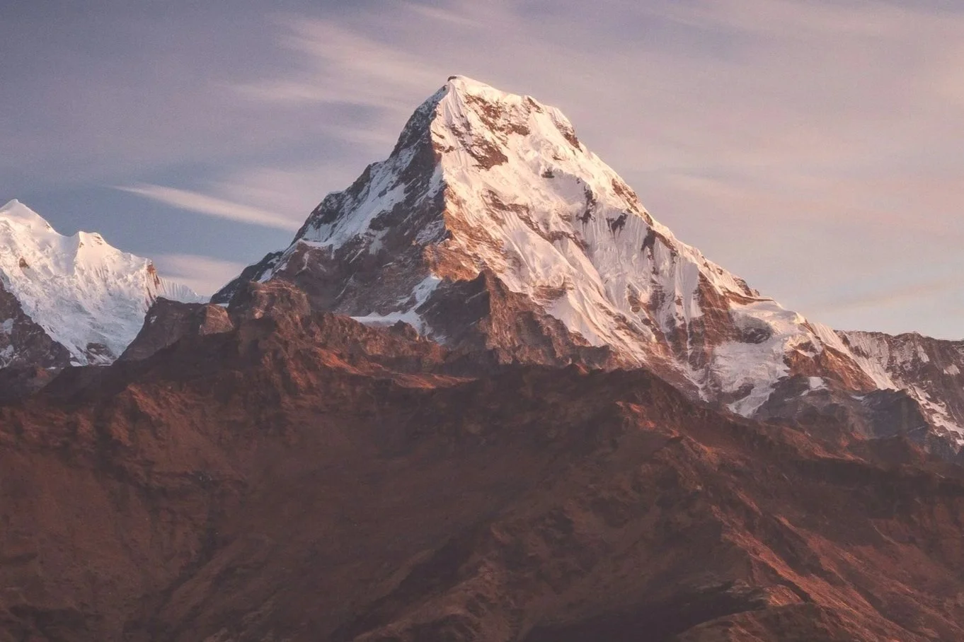 A snow-capped mountain peak, likely Mount Everest, at sunset with a partly cloudy sky.