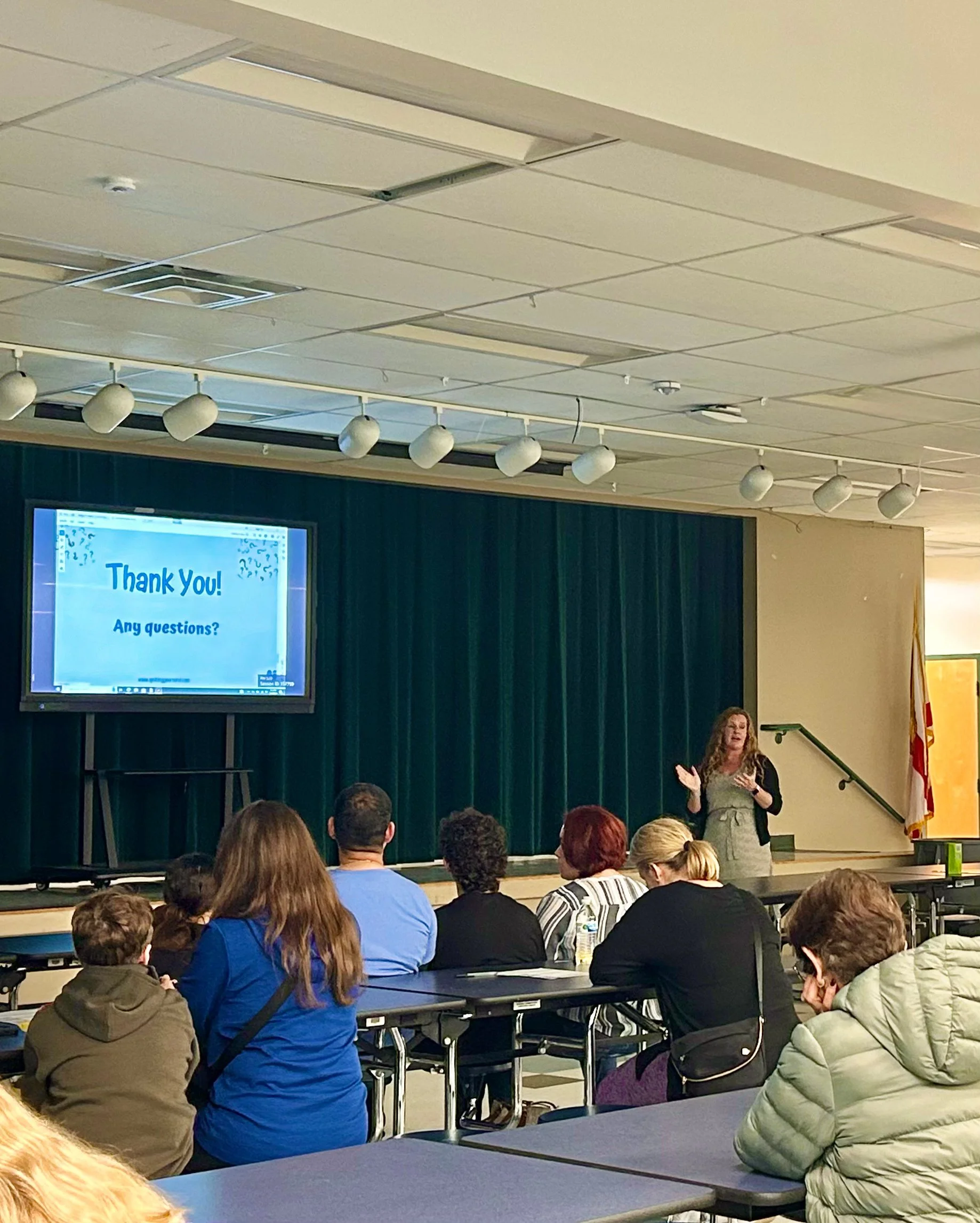 A woman is giving a presentation to an audience seated at tables in a conference room. There is a screen behind her displaying a message that says "Thank You!" and a question prompting for questions. The room has a ceiling with recessed lighting and a dark curtain backdrop.