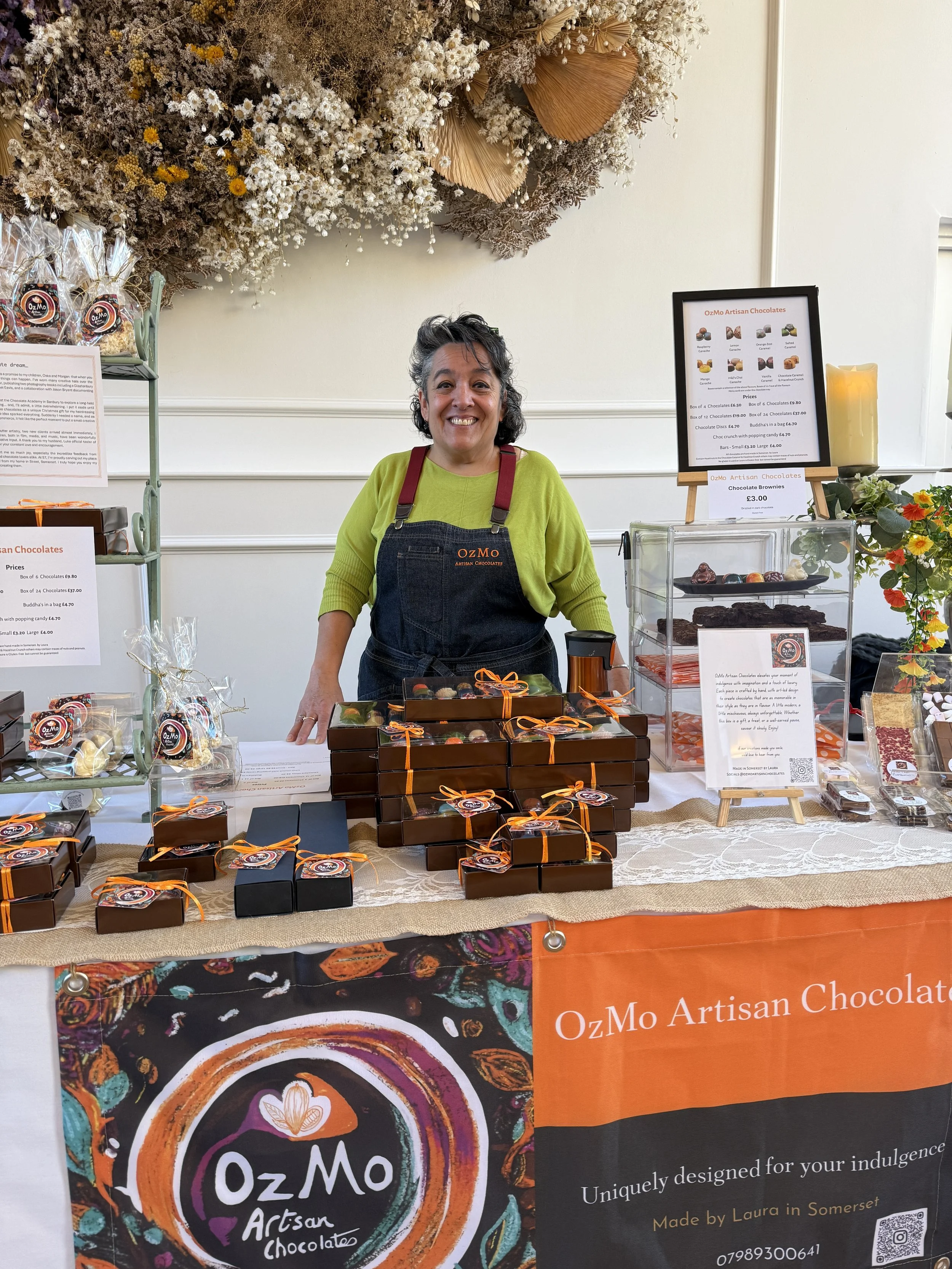 A woman wearing a green shirt and black apron with 'OzMo' logo stands behind a stall at a market, selling assorted artisan chocolates. The stall is decorated with a colorful banner featuring the OzMo logo, and the table displays wrapped chocolates, boxes, and a glass case with more chocolates. There are promotional signs and a large yellow candle on the side.