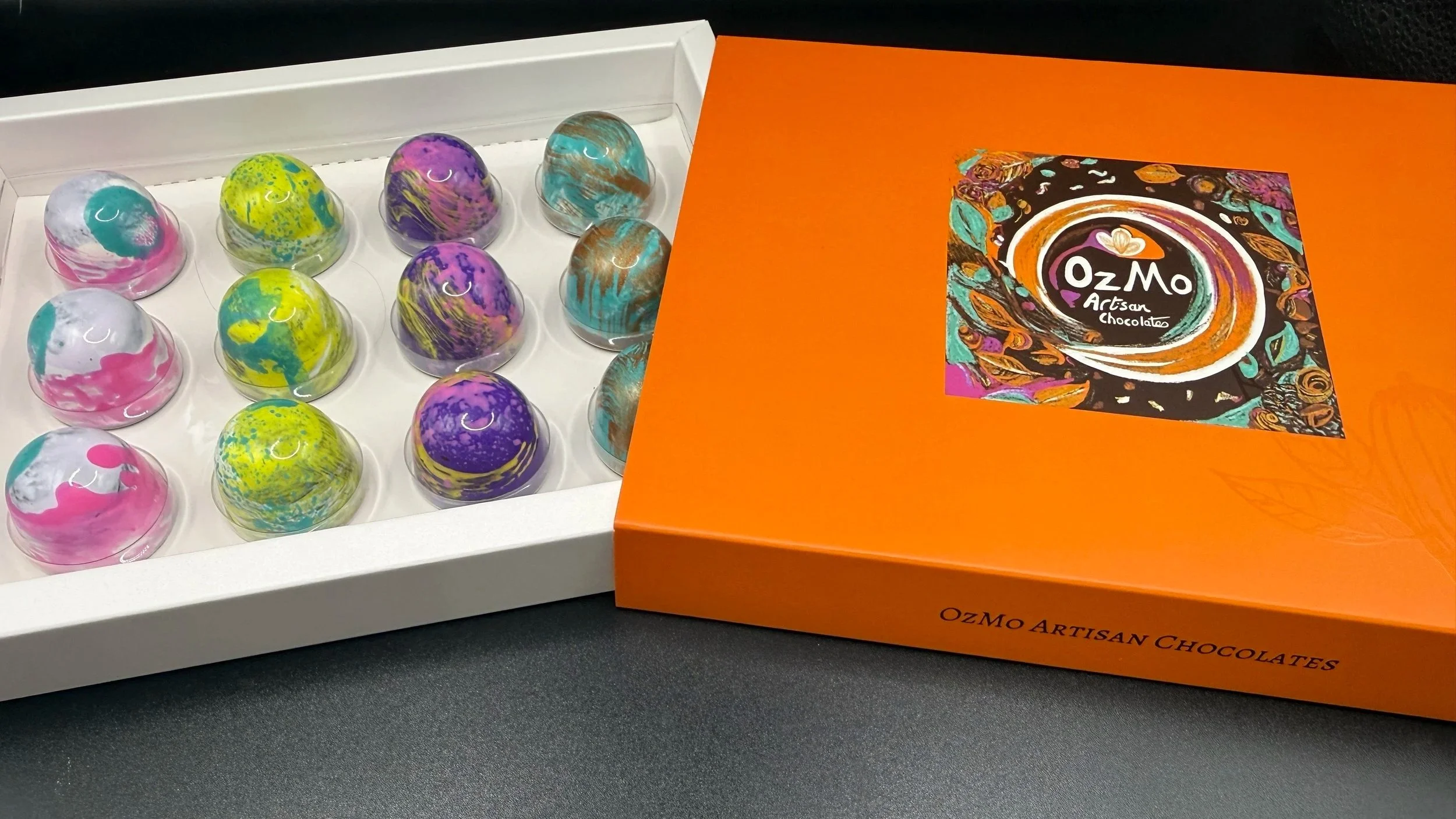 A box of 12 marbled chocolates with swirls of pink, yellow, purple, blue, and brown, next to an orange OzMo artisan chocolates box with colorful artistic design on the lid.