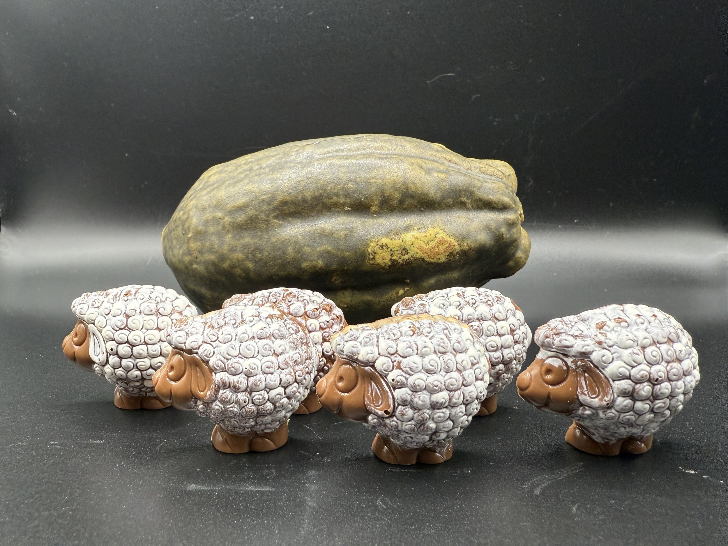 Five small artisan chocolate sheep with white curly wool and brown faces and legs, placed on a black surface with a dark background, and a ceramic coco pod.