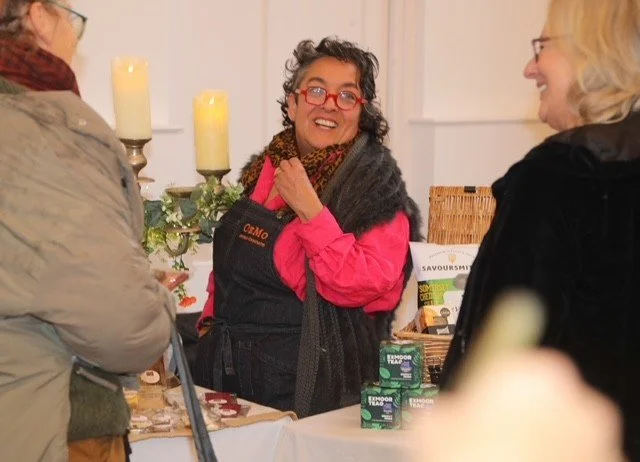 A woman with glasses and curly hair, wearing a pink shirt and apron, smiling and talking with two other women at a market or craft fair.