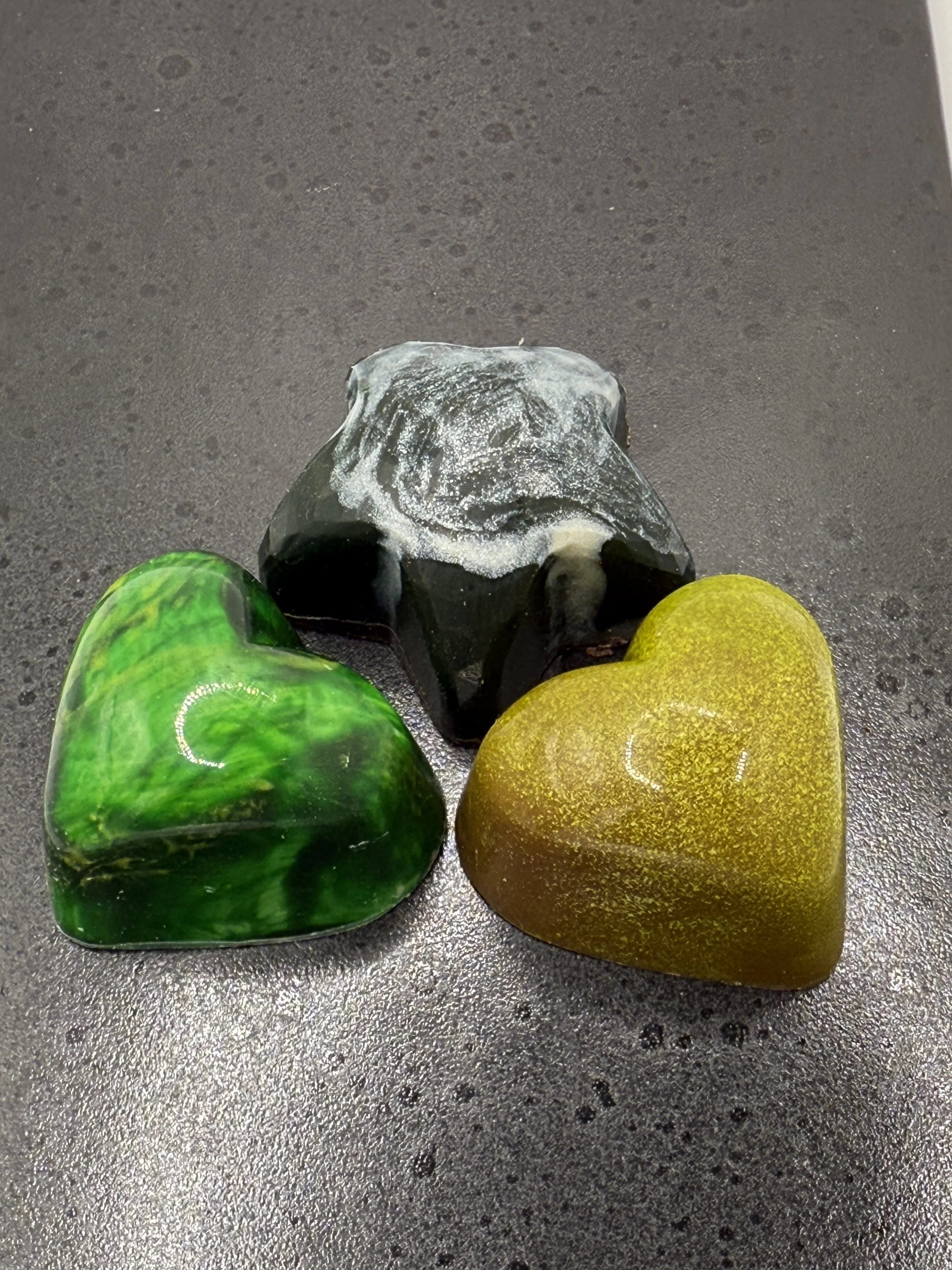Three polished heart-shaped artisan hand painted filled bonbon chocolates, one green with swirl pattern, one dark green with white streaks, and one yellow speckled, on a black speckled surface.