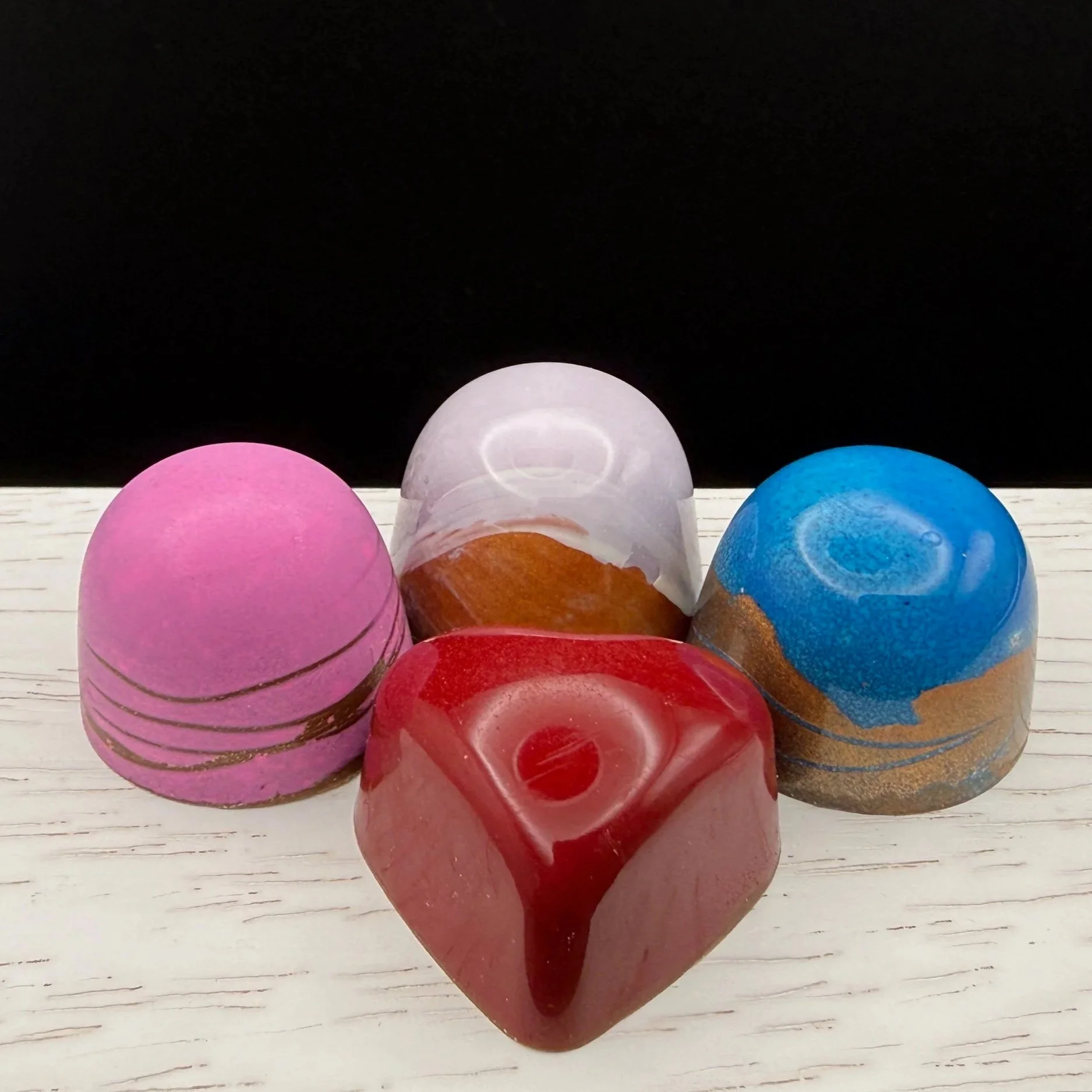 Four colorful chocolates arranged on a white wooden surface with a black background. The chocolates are pink with gold lines, clear with brown/amber accents, blue with gold and brown swirls, and red with a central hole and drippy design.