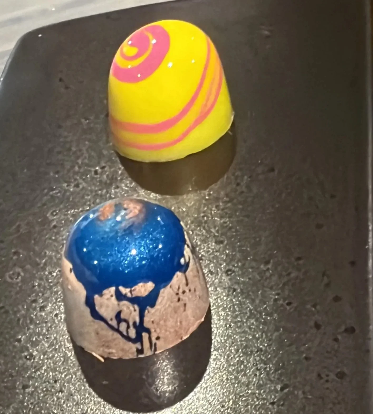 Decorative artisan handmade filled bonbon chocolates on a black surface, with one yellow candy featuring pink swirl designs and another blue candy with dark blue chocolate drips.