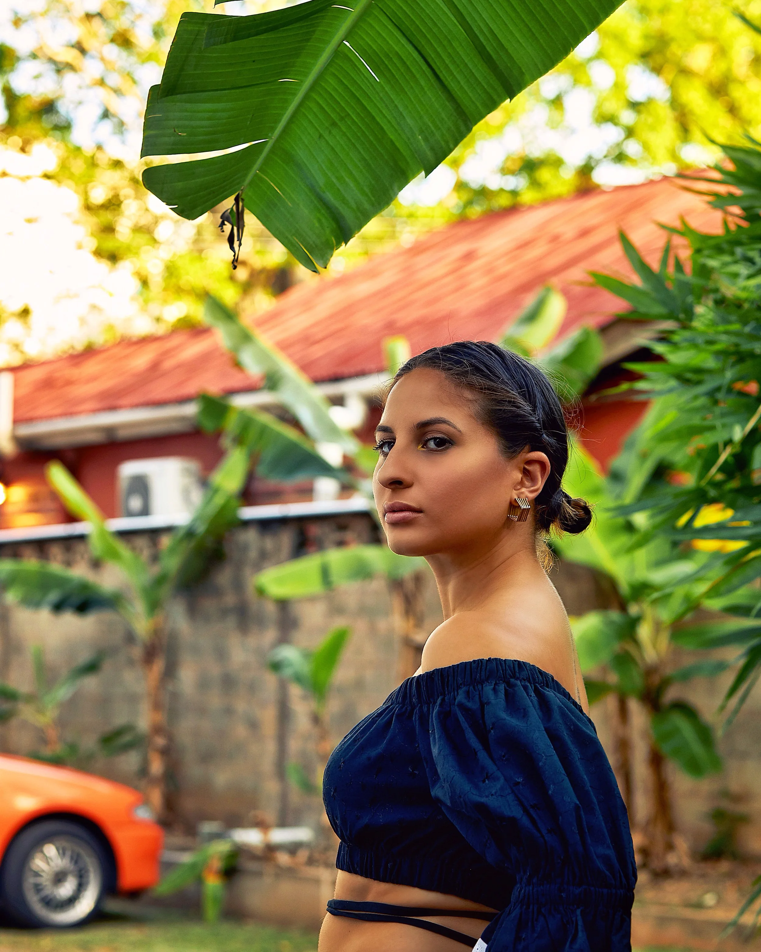 A young woman with dark hair styled in a low bun, wearing a dark off-the-shoulder top, standing outdoors among lush green plants, with a red-roofed building and a parked orange car in the background.