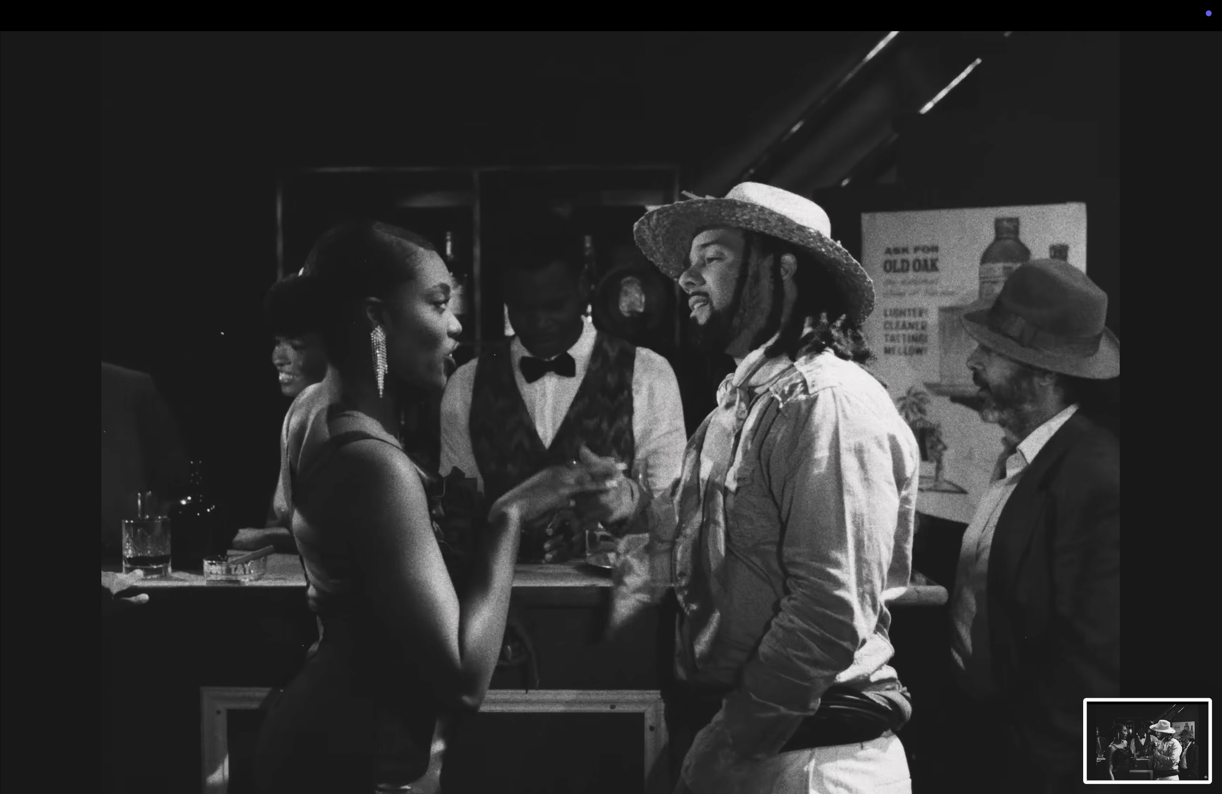 Black and white photo of a lively social scene with people dressed in vintage clothing, including a woman in a sleeveless dress and a man with a wide-brimmed hat and dreadlocks, engaging in conversation.