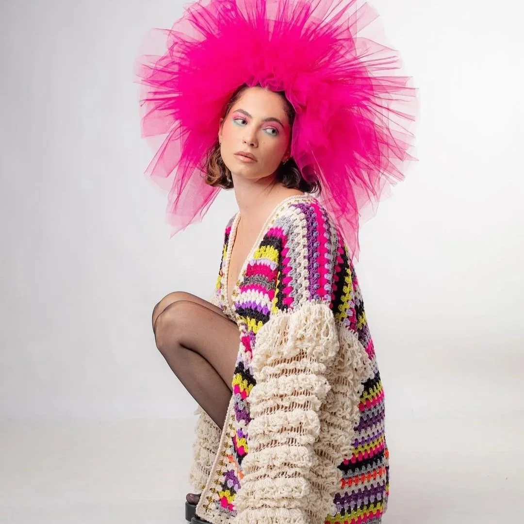 A woman wearing a large pink tulle hat, colorful crocheted sweater, black sheer tights, and high heels, posing against a white background.