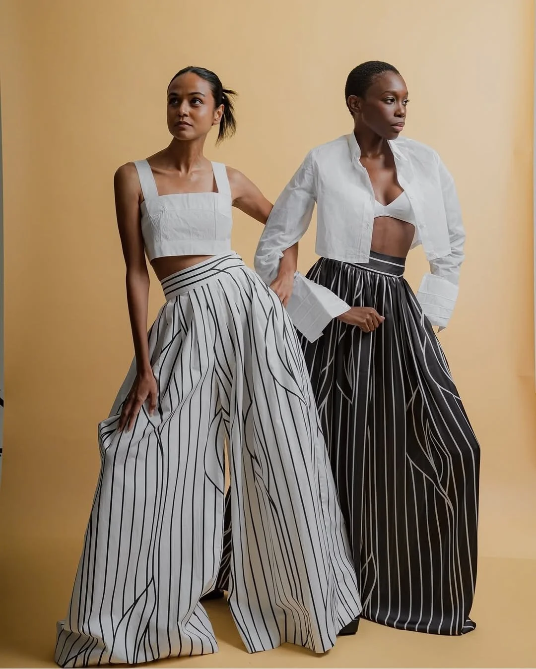 Two women posing against a beige background, wearing black and white striped wide-leg pants and white tops; one with a cropped top and the other with an unbuttoned shirt.