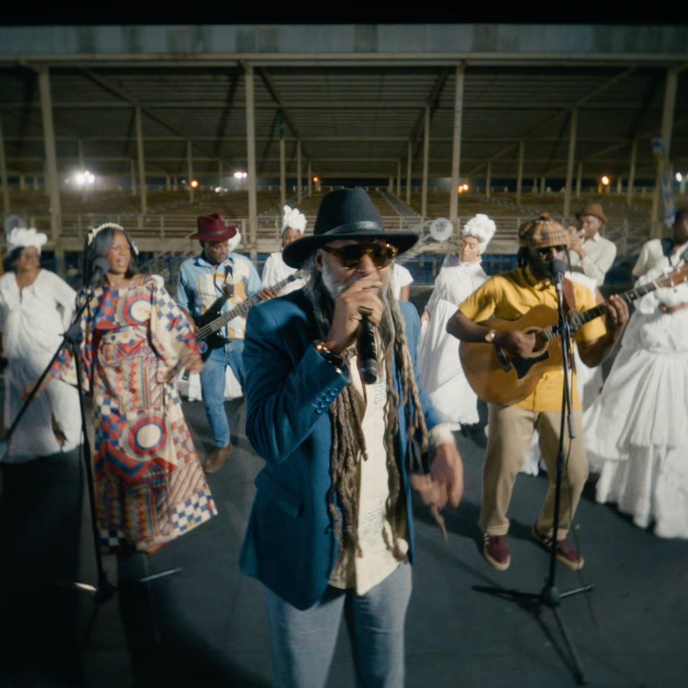 Musicians performing at an outdoor event, featuring a singer with dreadlocks, black hat, sunglasses, and a blue blazer, accompanied by a guitarist wearing a yellow shirt and tan pants.