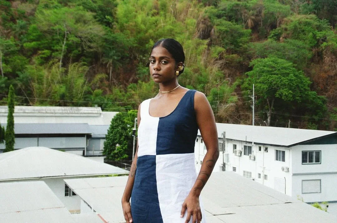 Young woman standing outdoors with green trees and white buildings in the background, wearing a sleeveless dress with blue and white color blocks and gold jewelry.