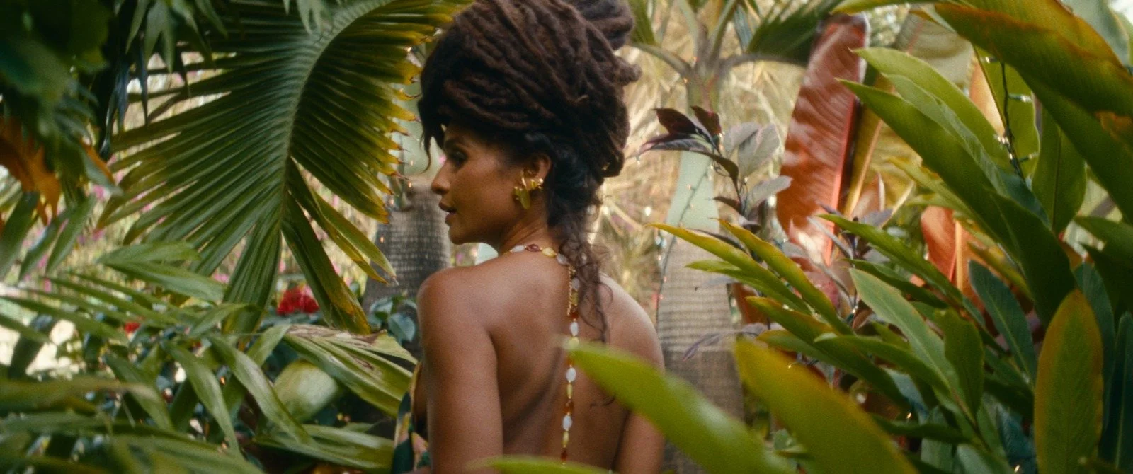 A woman with dreadlocks and gold accessories stands among lush tropical plants and palm trees in a jungle setting.