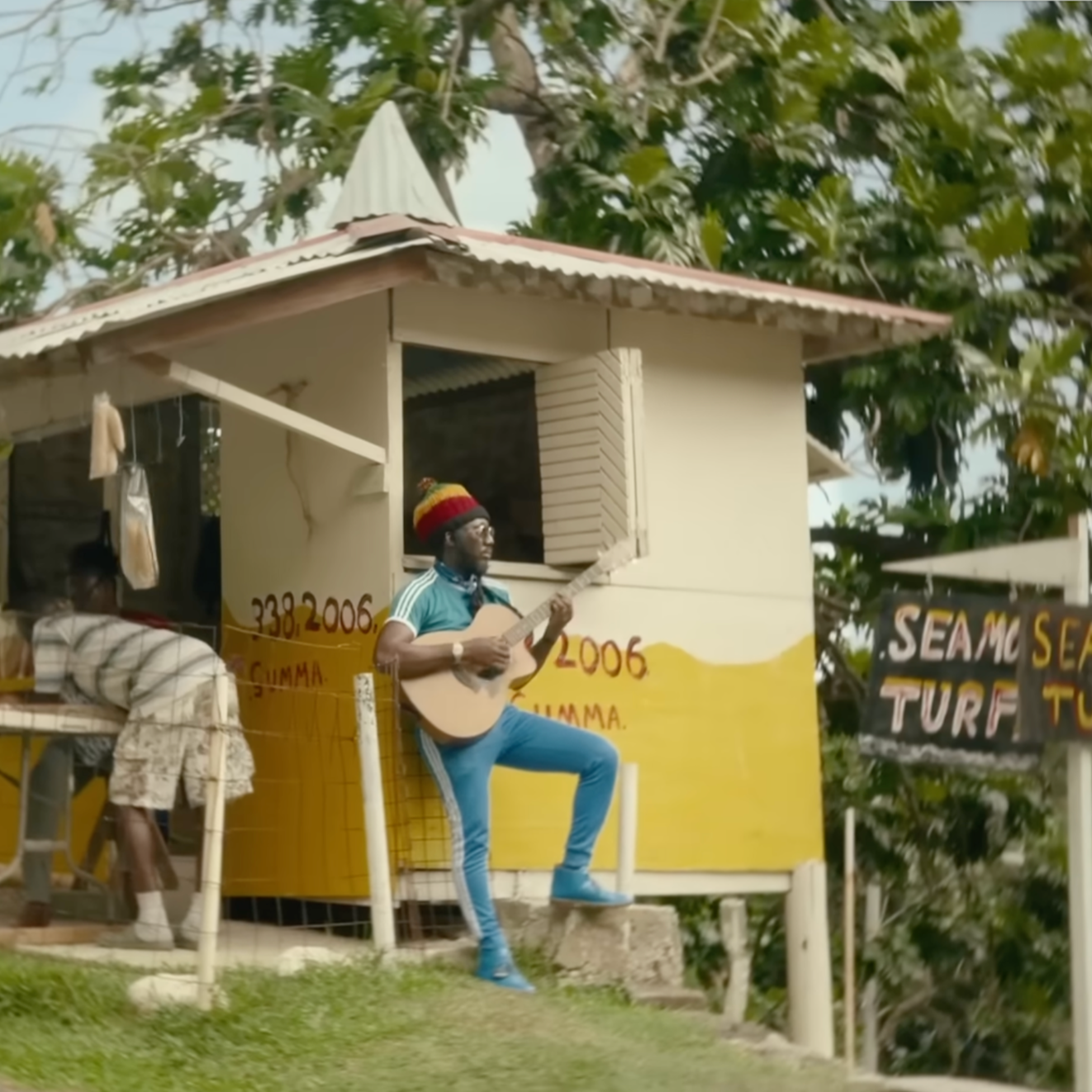 Man playing guitar on a small elevated yellow building with window shutters, with a person sitting nearby, surrounded by trees, and a sign nearby that reads 'SEAMO' and 'TURF'.