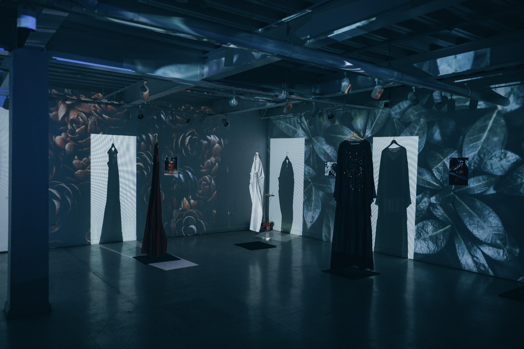 Art installation with dresses hanging on white display boards, shadows and light effects on the wall, surrounded by floral patterns projected onto the walls and ceiling.