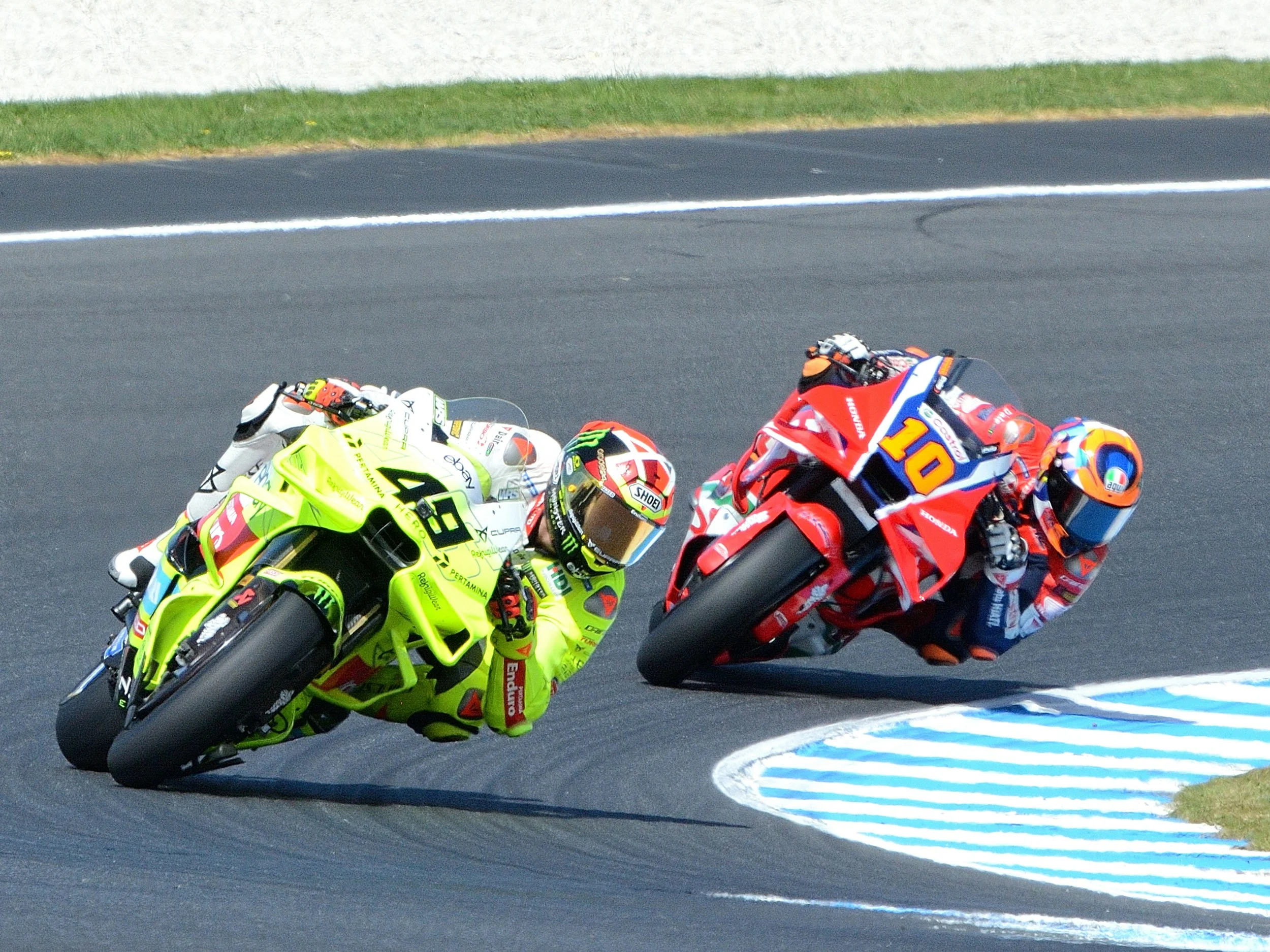 Two motorcycle racers leaning into a turn on a racetrack, one in bright yellow and the other in red, with a blue and white striped curb on the inside of the curve.