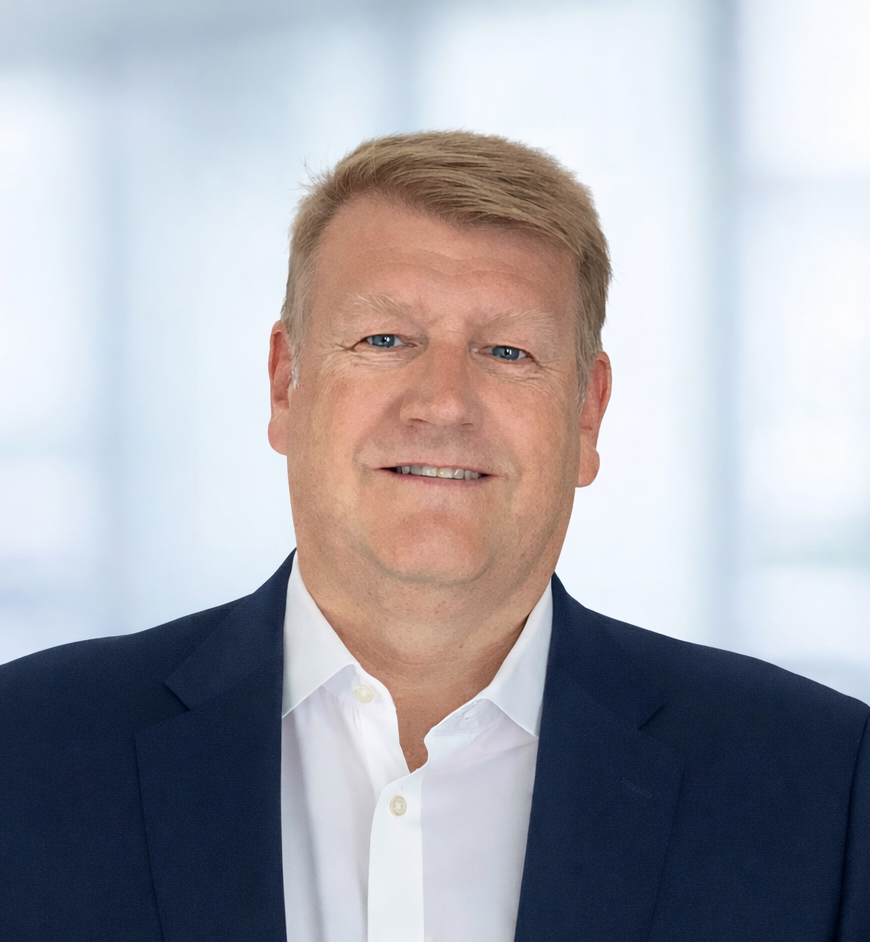 A middle-aged man with blonde hair wearing a dark suit and white shirt, smiling, against a light, out-of-focus background.