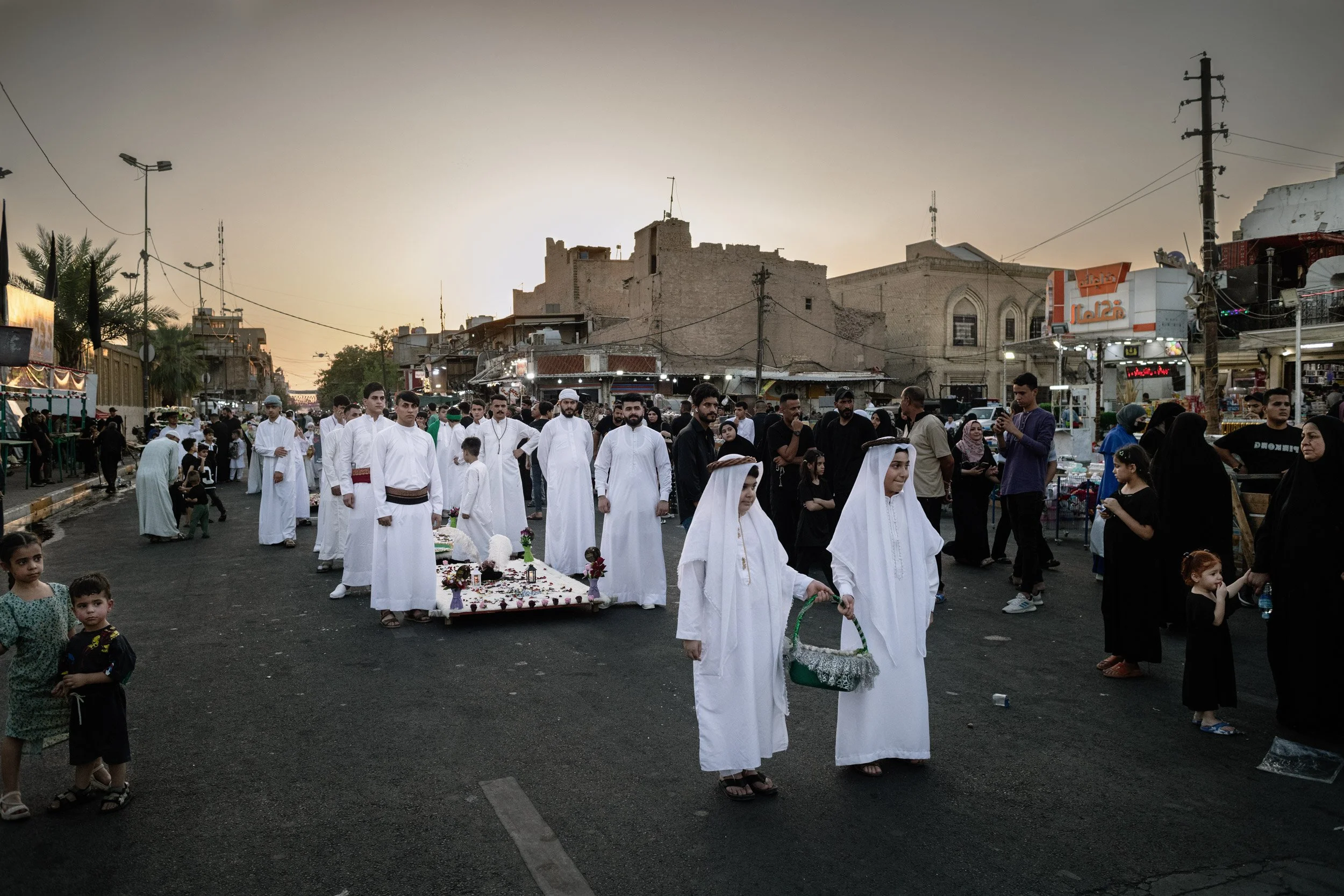 Un groupe de personnes vêtues de blanc participent à une procession ou cérémonie dans une rue animée au coucher du soleil, entourée de bâtiments traditionnels et de stands de marché. Bagdad, Iraq