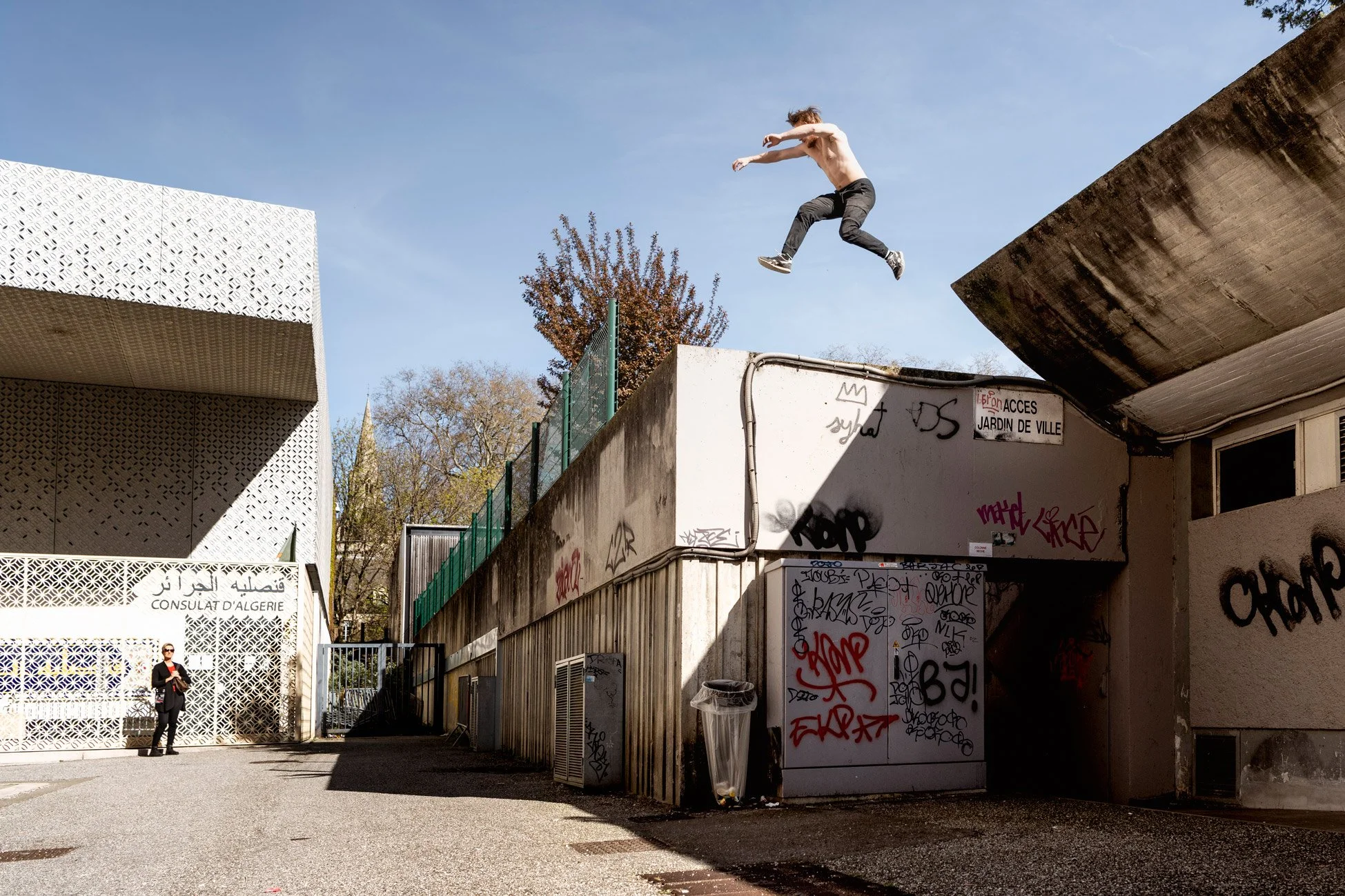 Un jeune homme en short saute d'une hauteur sur un mur en milieu urbain. À gauche, une femme en costume noir observe. Les murs sont recouverts de graffitis, et la scène se déroule en plein jour sous un ciel ensoleillé.